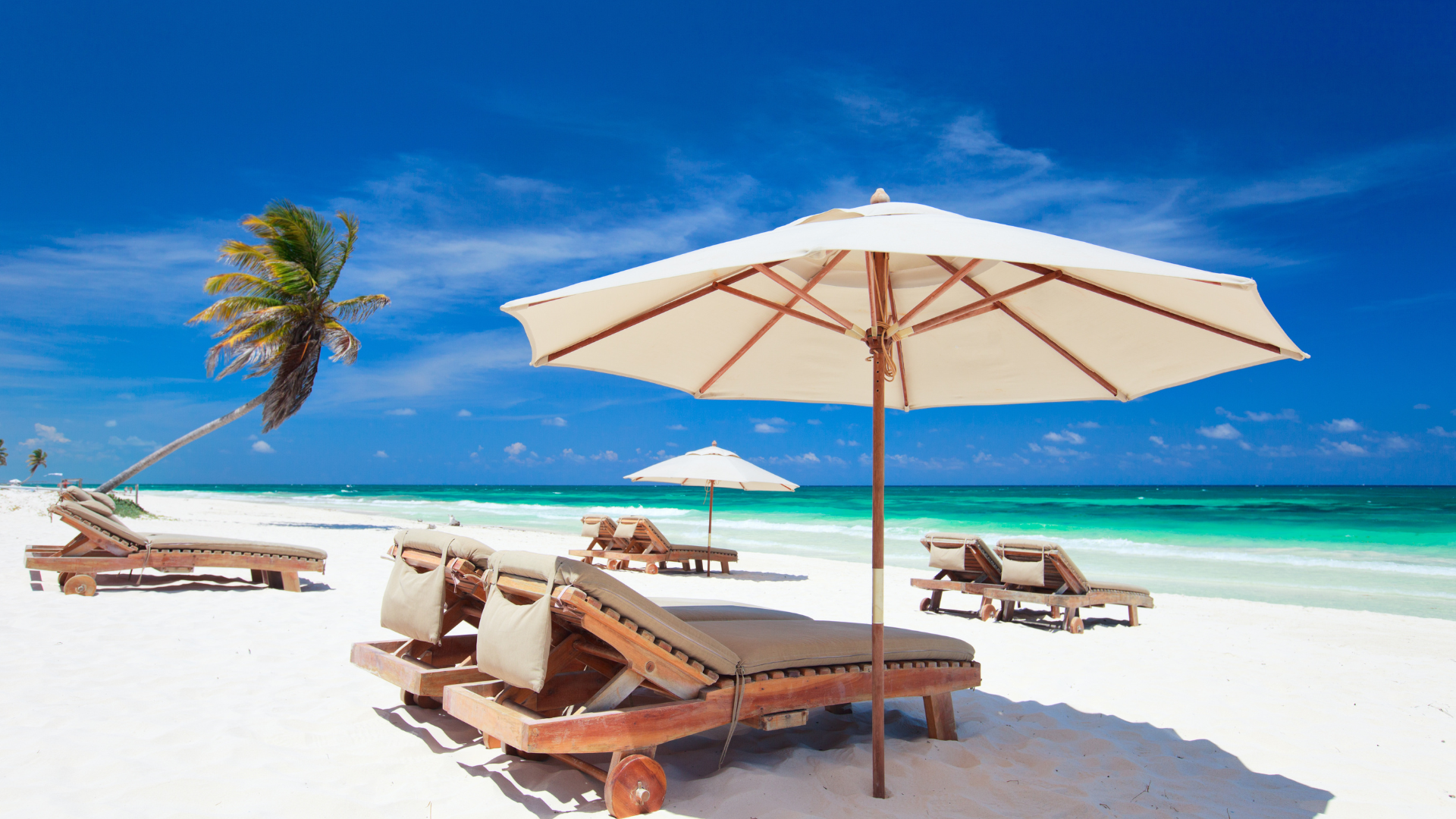 Empty beach with lounge chairs, large white beach umbrellas, a leaning palm tree, white sand, and turquoise ocean under a blue sky.