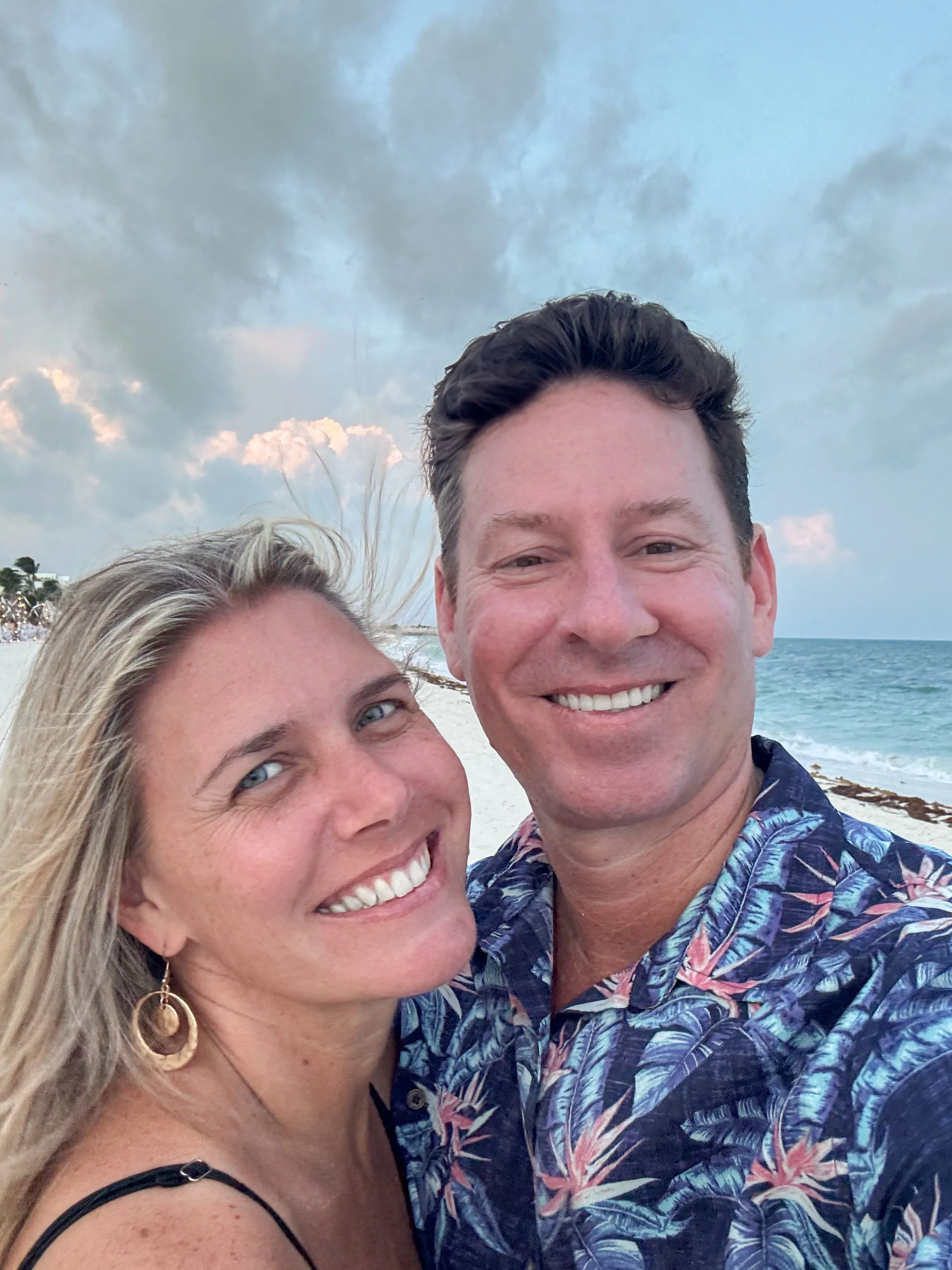 A smiling man and woman taking a selfie on a beach at dusk with the and Caribbean Sea in the background.