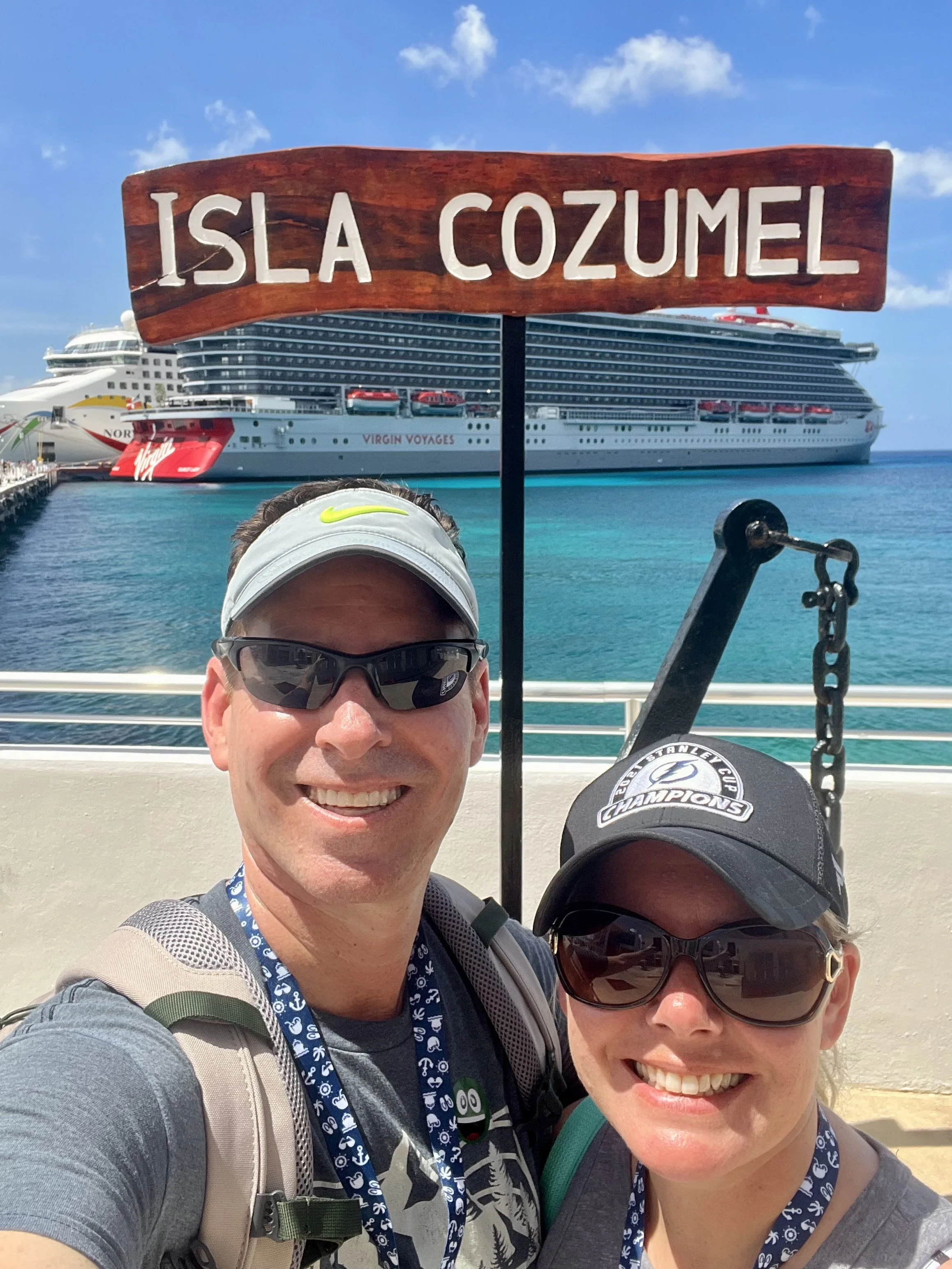 Two smiling people taking a selfie at Isla Cozumel with a cruise ship in the background on a sunny day.