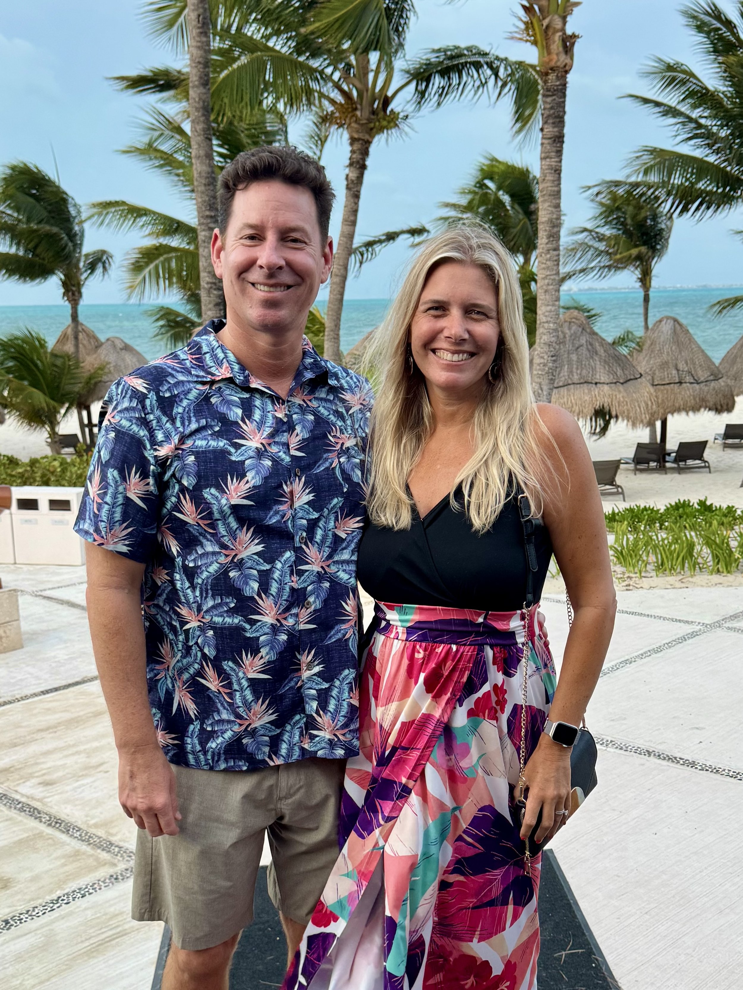 A smiling man and woman standing close together outdoors with a beach scene in the background, including palm trees, ocean, and thatched umbrellas.