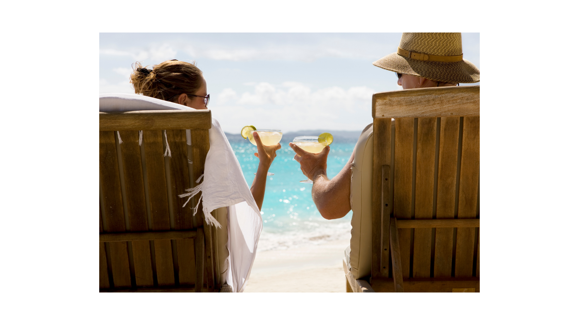 A couple sitting on beach chairs by the ocean, holding tropical drinks with lime slices, enjoying a sunny day.