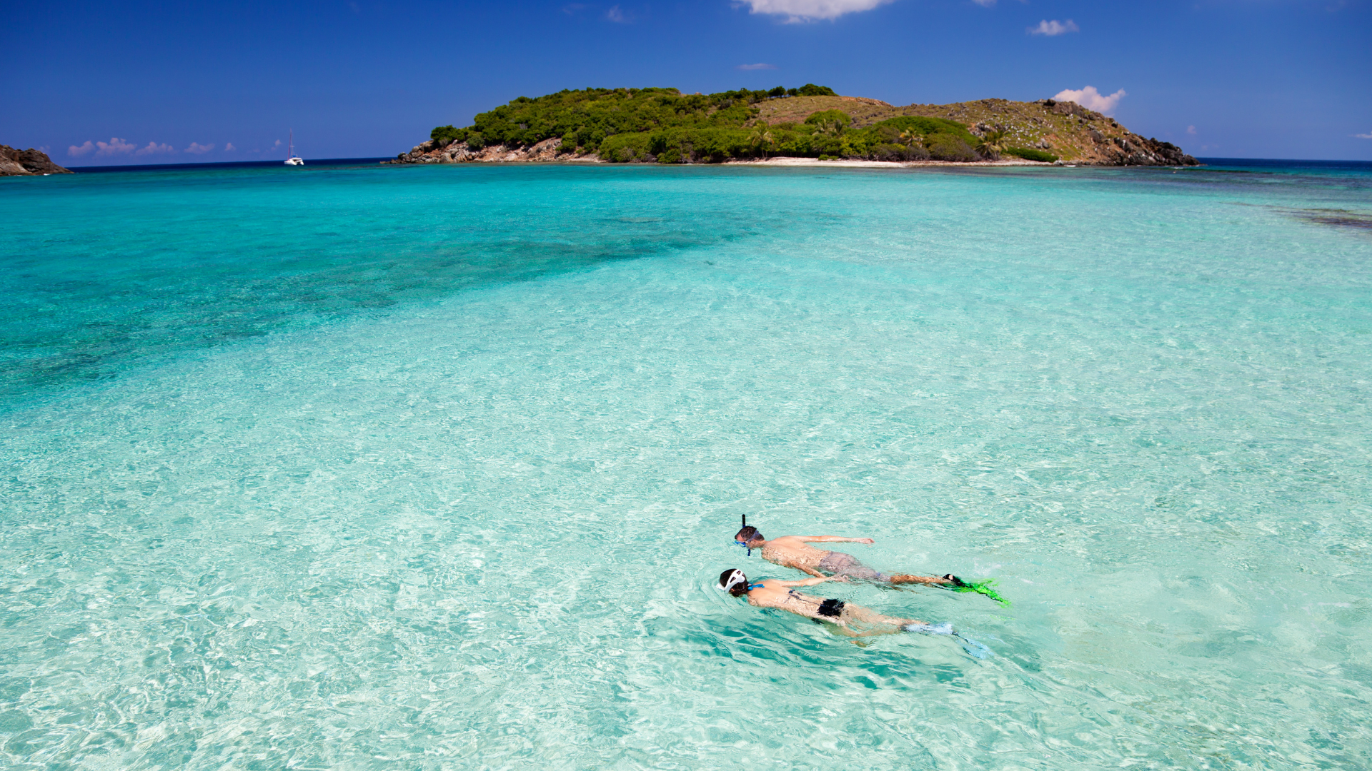 Two people snorkeling in clear turquoise water near a sandy shore with a green, tree-covered island in the background under a partly cloudy sky.