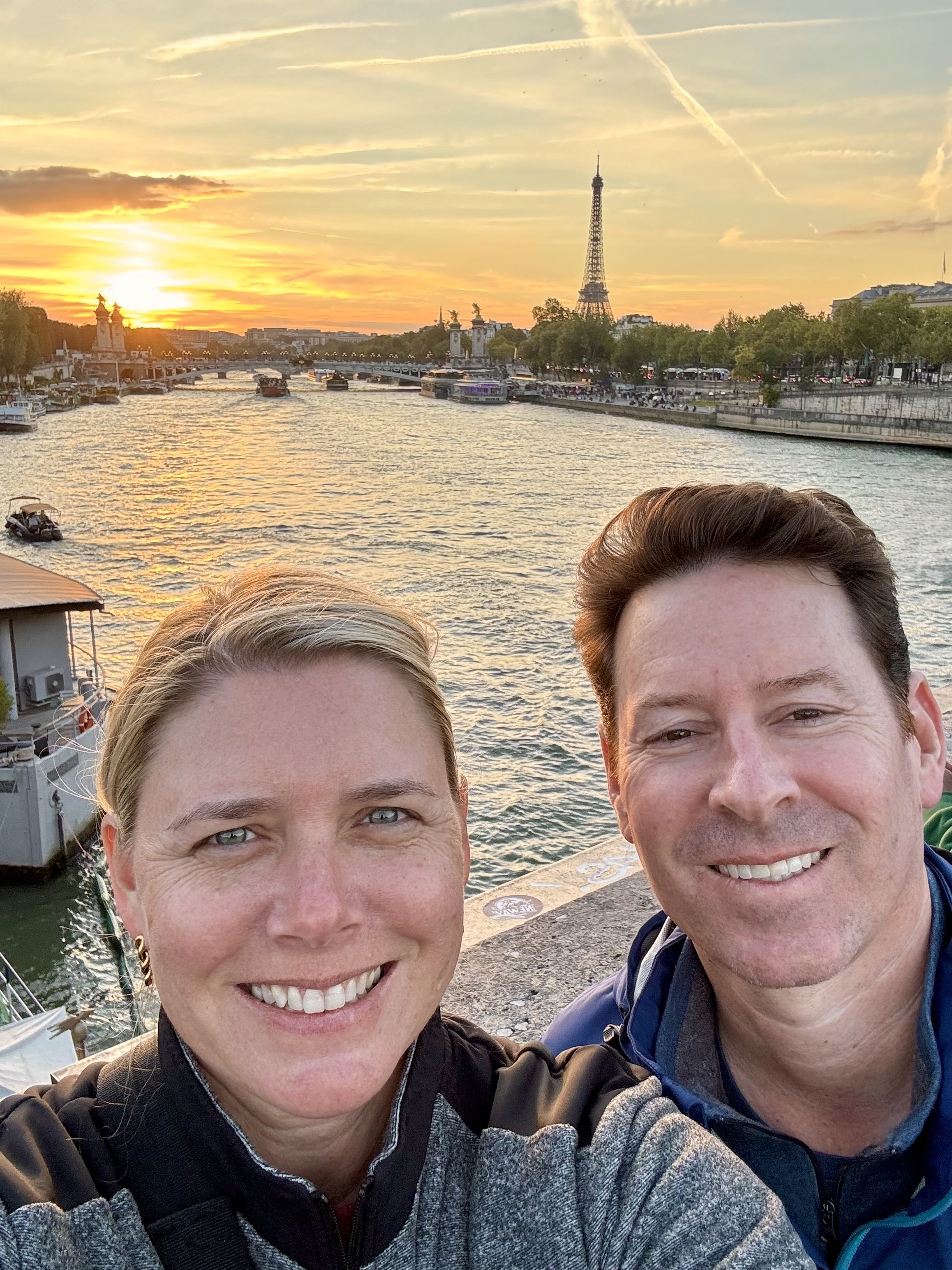 Two people taking a selfie by the river at sunset, with the Eiffel Tower visible in the background in Paris, France.