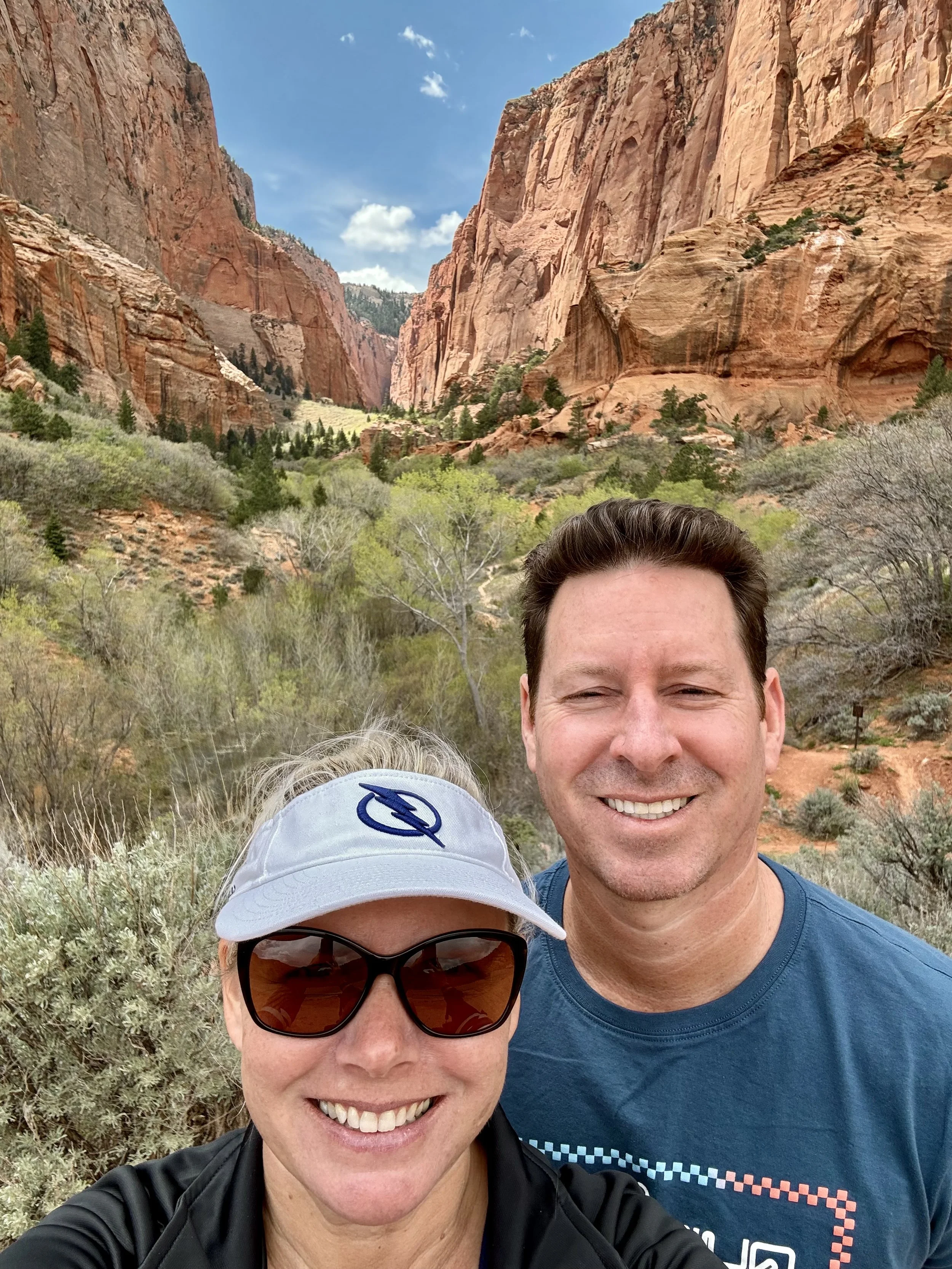 Two smiling people taking a selfie in a scenic canyon with tall red rock formations and greenery in the background.