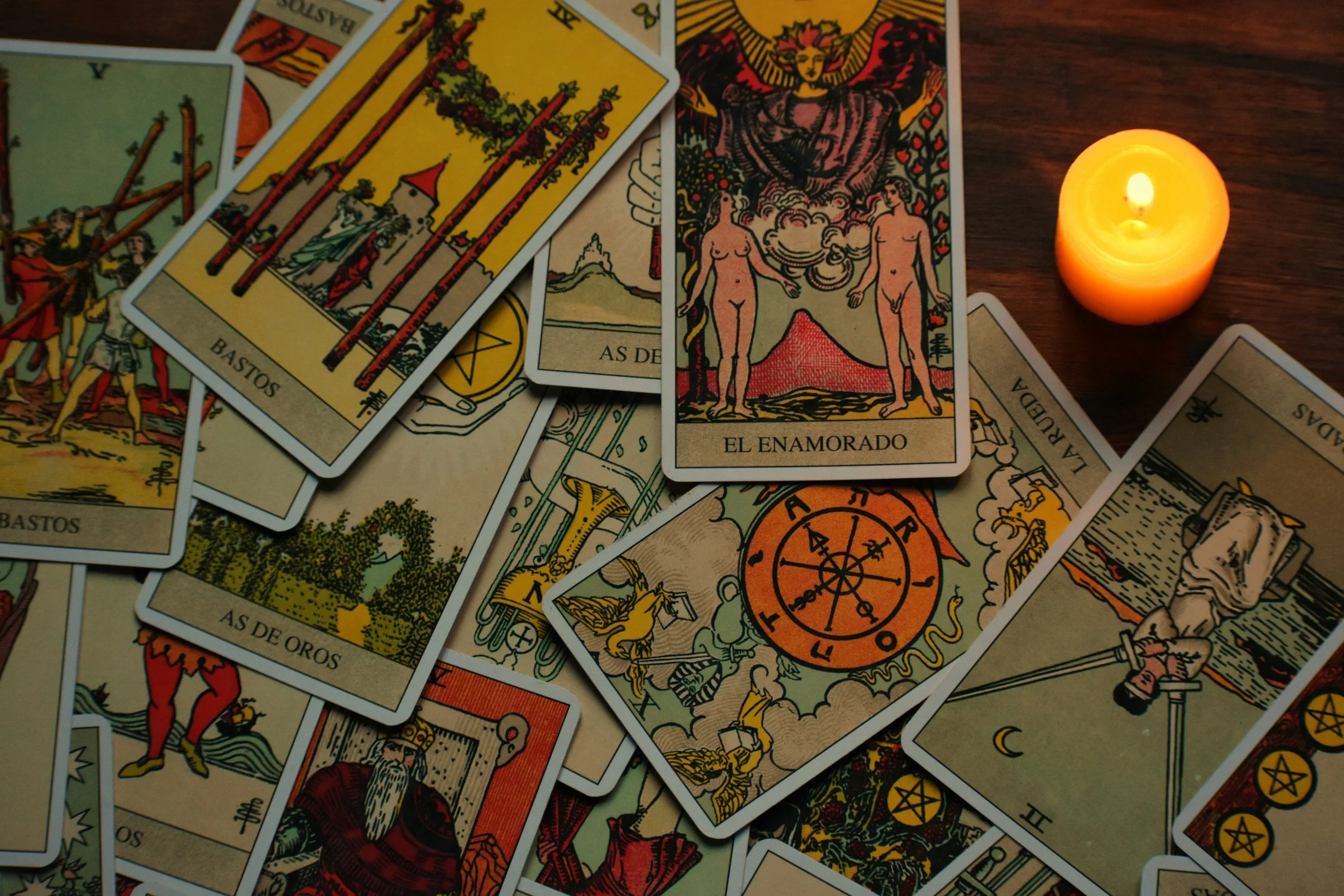 A spread of Tarot cards on a wooden surface, illuminated by a small lit yellow candle, with some cards overlapping each other.
