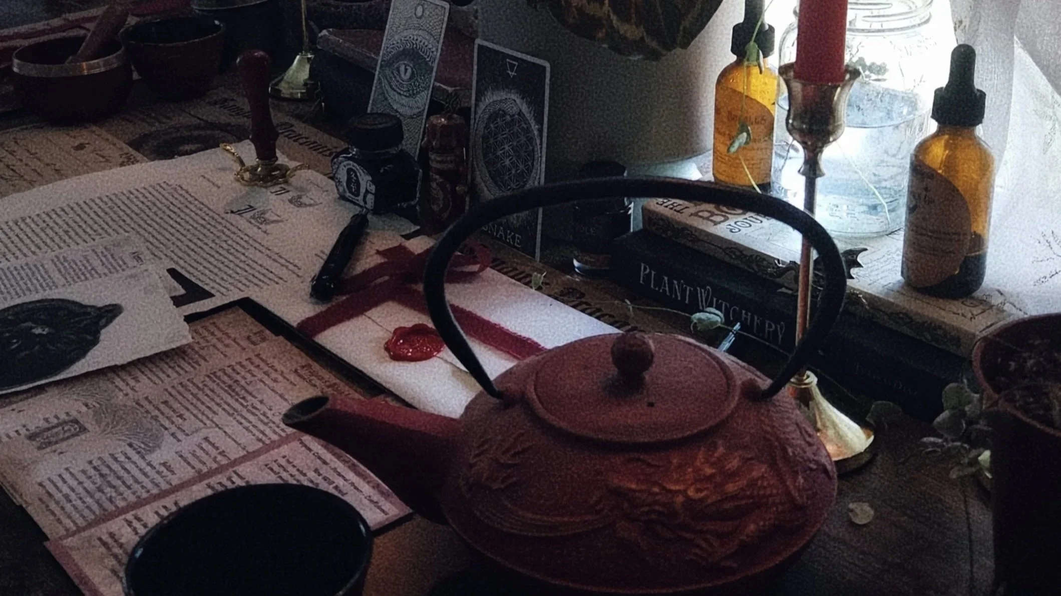 A tabletop filled with various objects including candles, bottles, a teapot, a stack of cards, and newspapers, with a window in the background letting in light.
