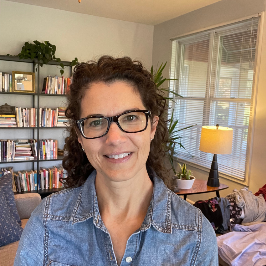 A woman with curly brown hair wearing glasses and a denim shirt, smiling, in a living room with a bookshelf, plants, a window with blinds, a lamp, and some clothes on a couch.