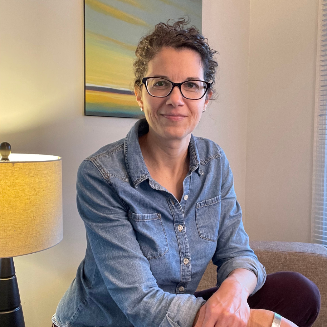 A woman with short curly hair and glasses sitting on a sofa in a living room, wearing a denim shirt, with a painting and a lamp in the background.