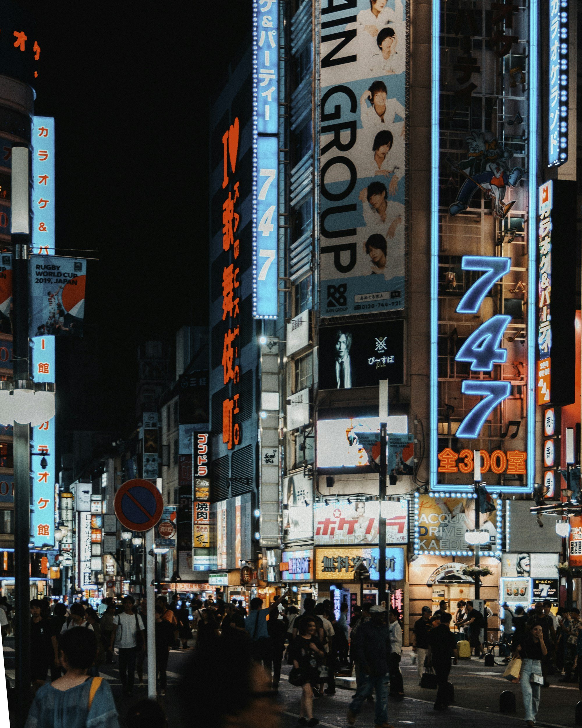 Une scène nocturne dans un quartier animé de Tokyo, avec de nombreux néons lumineux en japonais, des panneaux publicitaires, une grande foule de personnes marchant dans la rue, des taxis et des véhicules, et des gratte-ciel illuminés en arrière-plan.