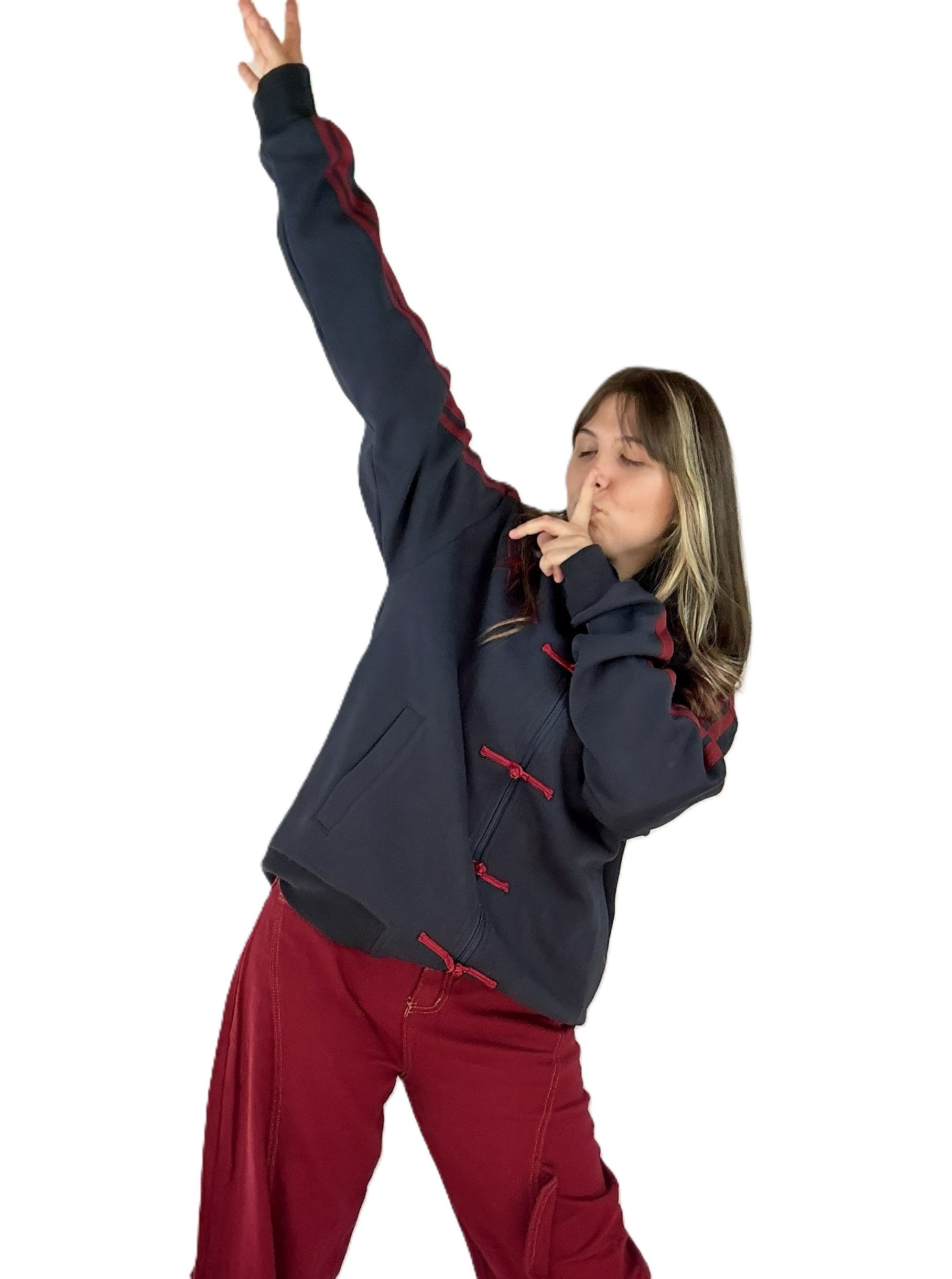 Une femme porte une veste noire avec des détails rouges et un pantalon rouge, en train de faire un geste de silence avec son index sur ses lèvres.