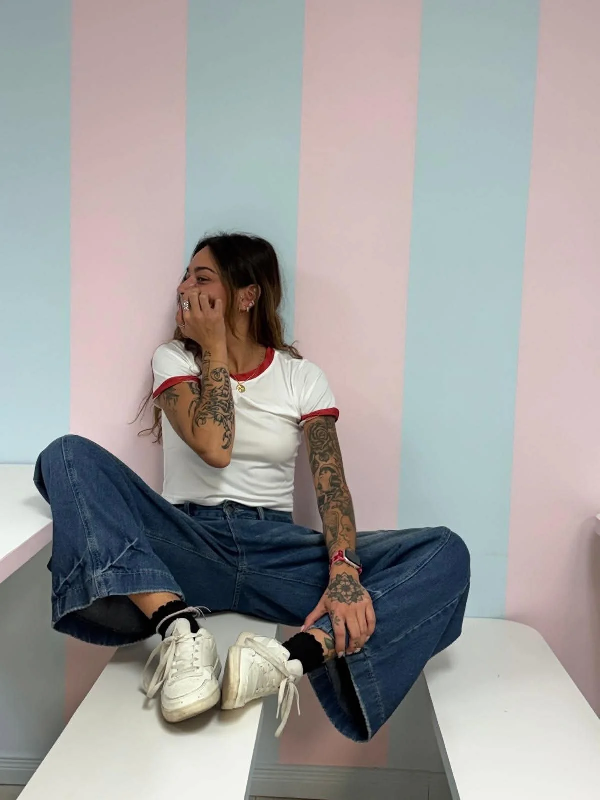 Jeune femme tatouée assise sur une table, riant et couvrant sa bouche, portant un t-shirt blanc à bord rouge, des jeans larges, et des baskets blanches, dans une pièce aux murs à rayures pastel.