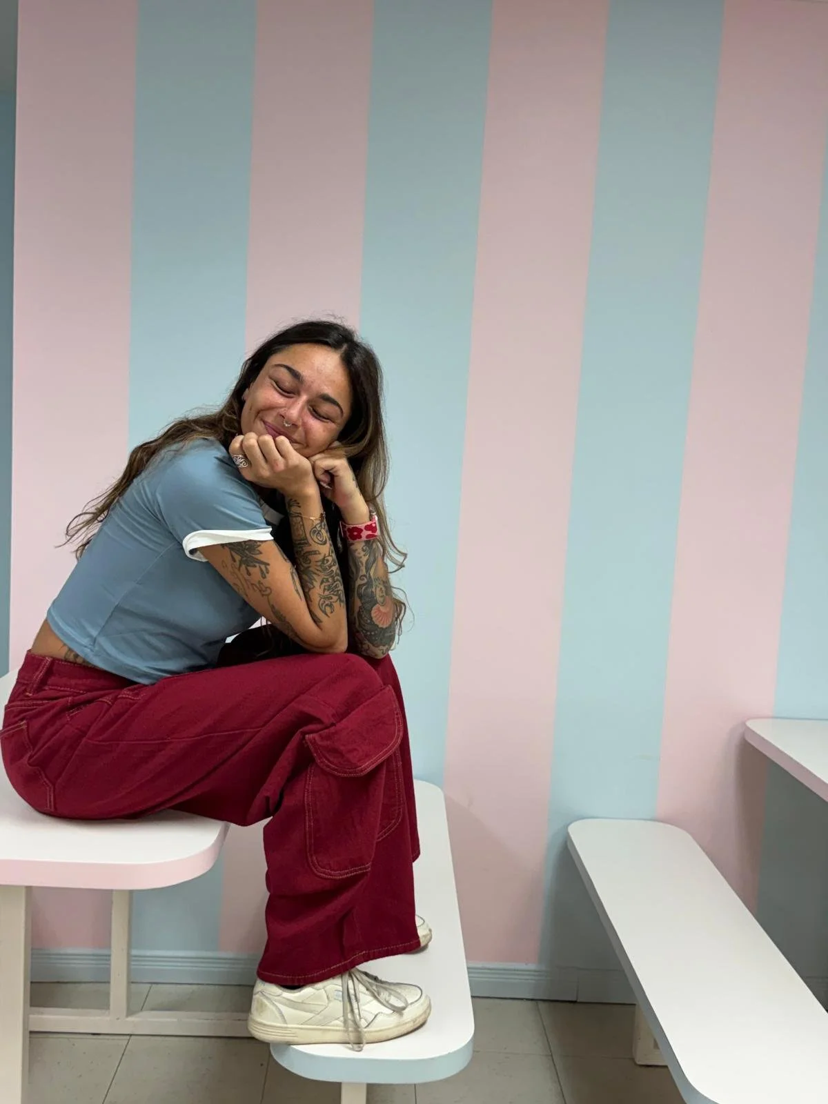 Une jeune femme assise sur une table, souriant et avec les yeux fermés, dans une pièce avec un mur à rayures pastel. Elle porte un t-shirt bleu, un pantalon rouge et des baskets blanches, et a des tatouages visibles sur ses bras.