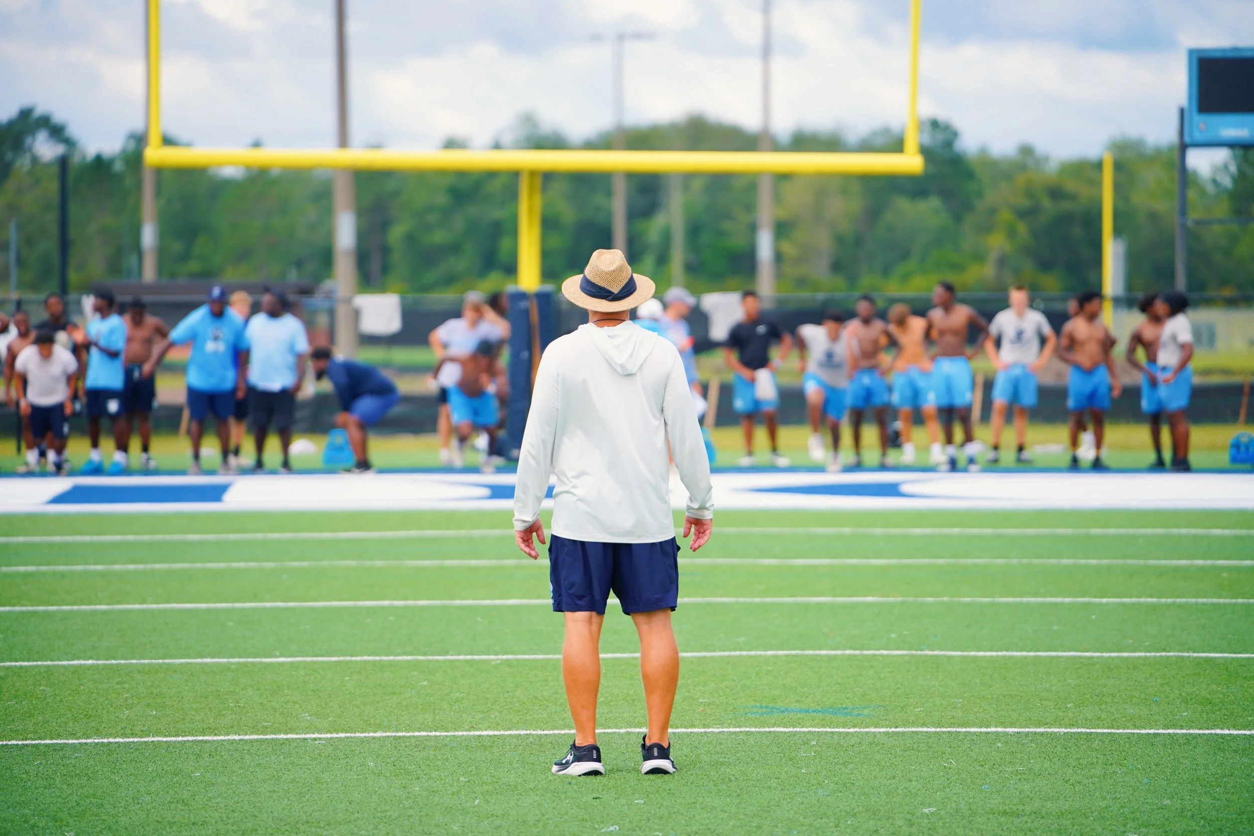Coach standing on football field observing players during practice.