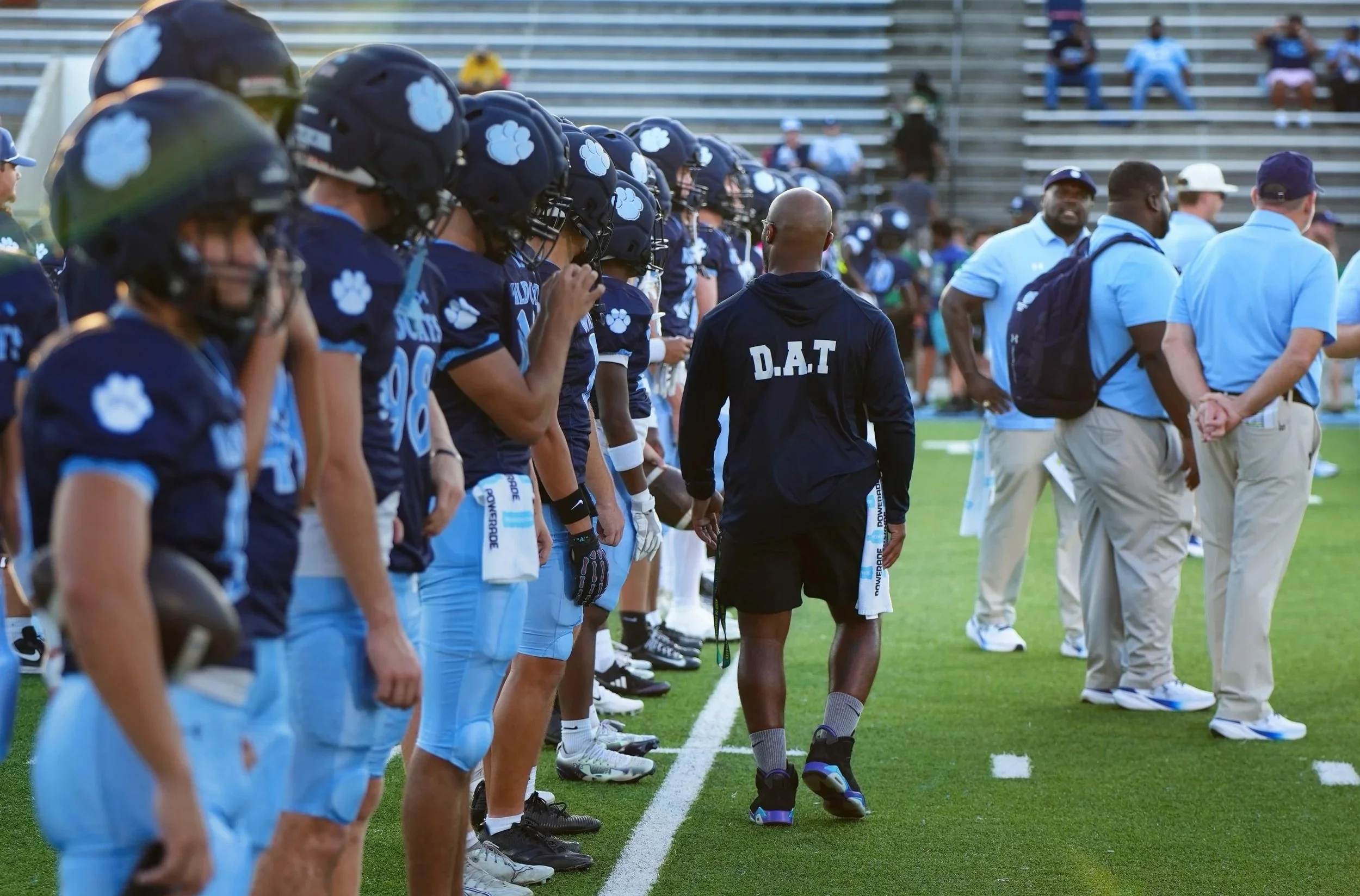 Football players in dark uniforms with blue accents and helmets featuring a cloud emblem, lined up for a game on the field, with coaches and officials nearby.