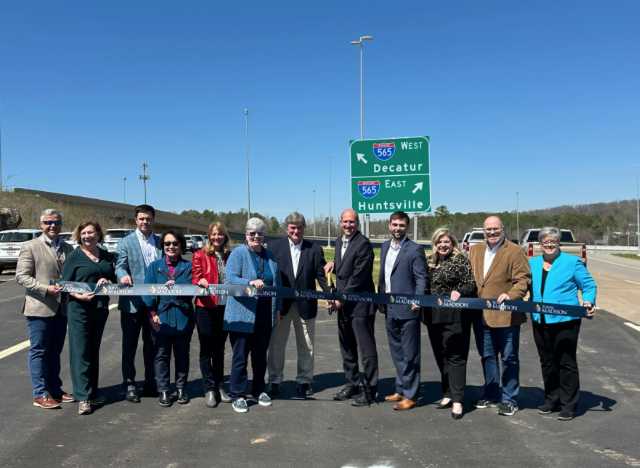 Group of people at a ribbon-cutting ceremony on a newly paved highway with a sign for Interstate 565 pointing east towards Huntsville and west towards Decatur.