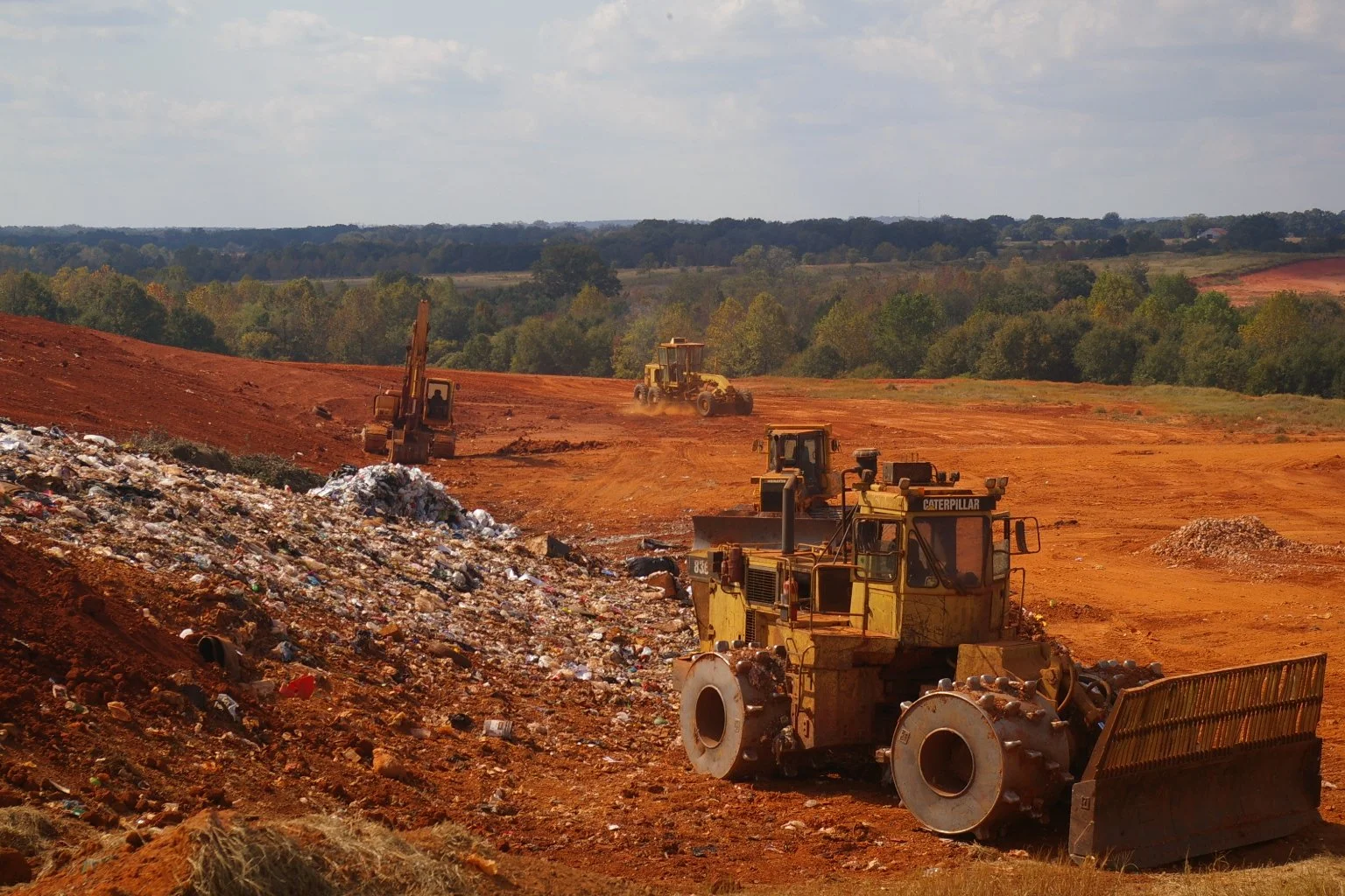 Construction site with multiple bulldozers and excavators on red dirt, with a background of green trees and cloudy sky.