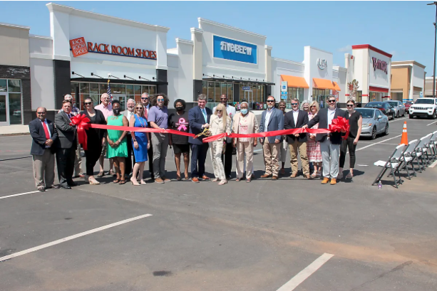 Group of people at a ribbon-cutting ceremony outside a shopping center with stores like Five Below and X Nonsense.
