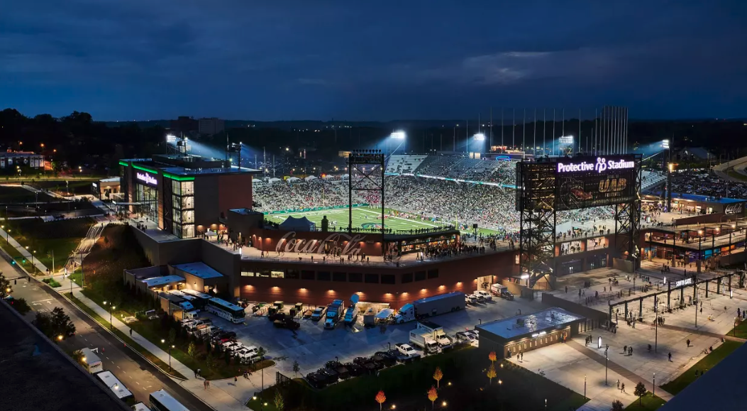Night view of Protective Stadium filled with spectators, illuminated with bright stadium lights, with a parking lot and surrounding buildings visible.