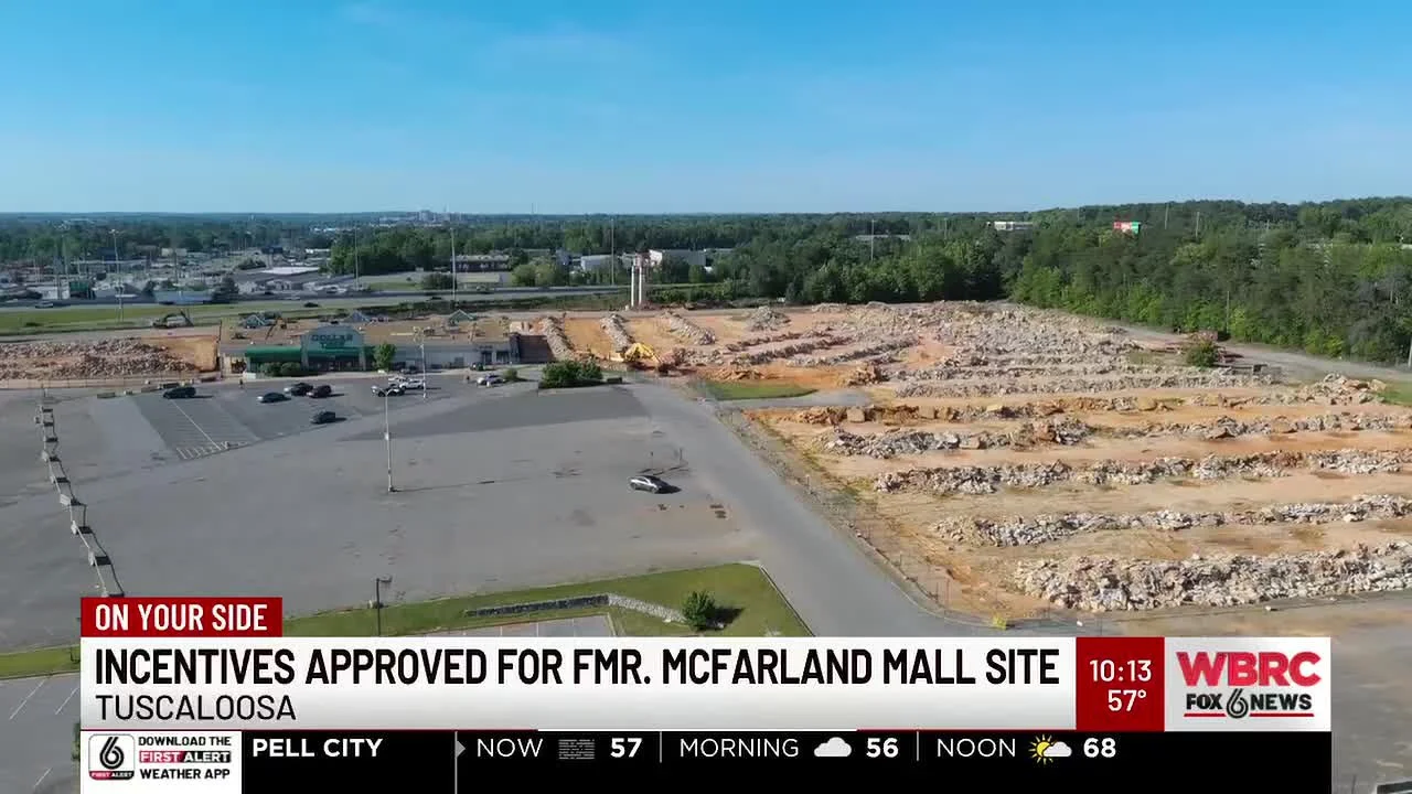 Aerial view of the old McFarland Mall site, with cleared land and construction equipment, parking lot, and surrounding trees, under a blue sky.