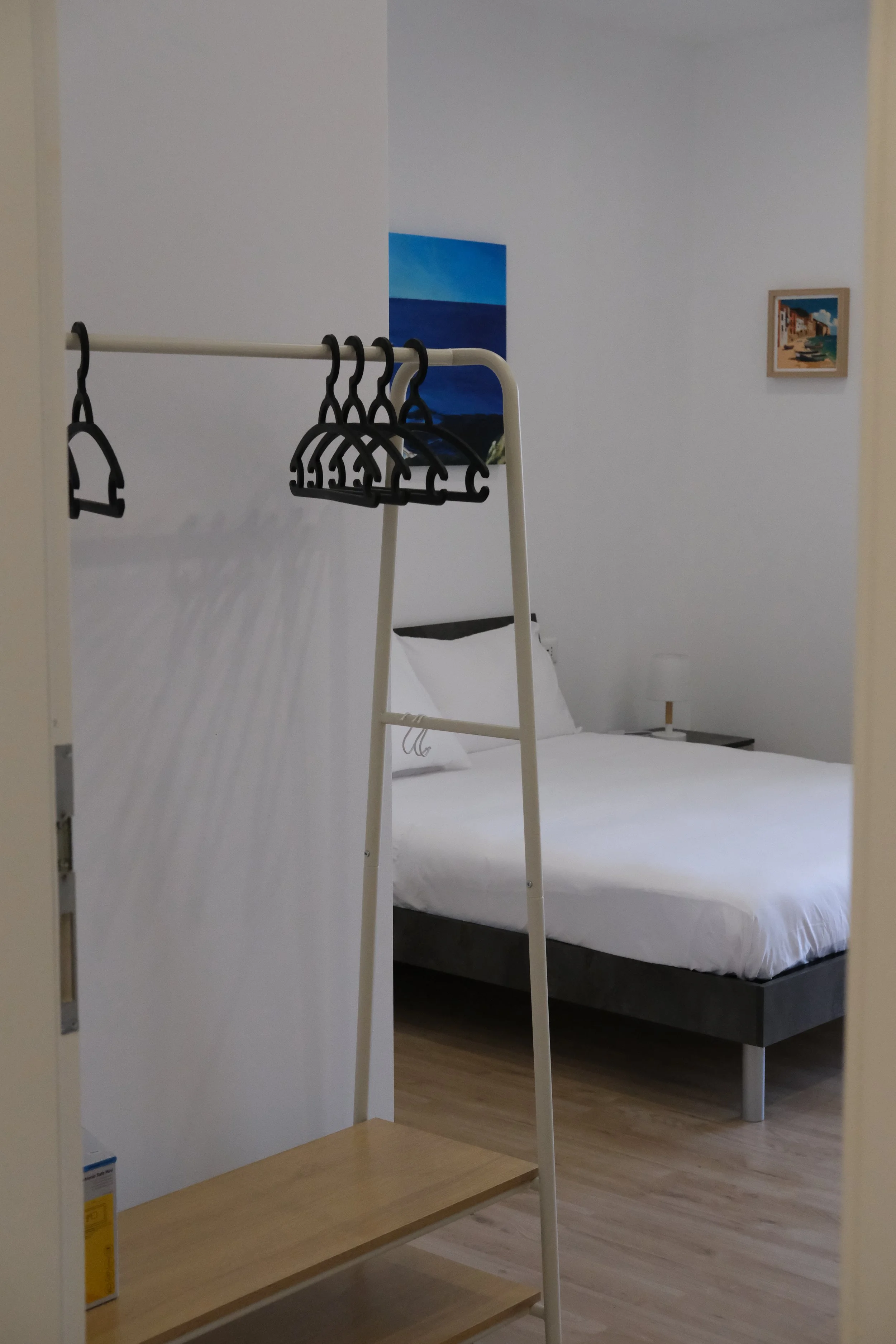 Camera da letto minimalista con letto matrimoniale bianco, quadri sulla parete, e scaffale di legno con portabiti vuoto.