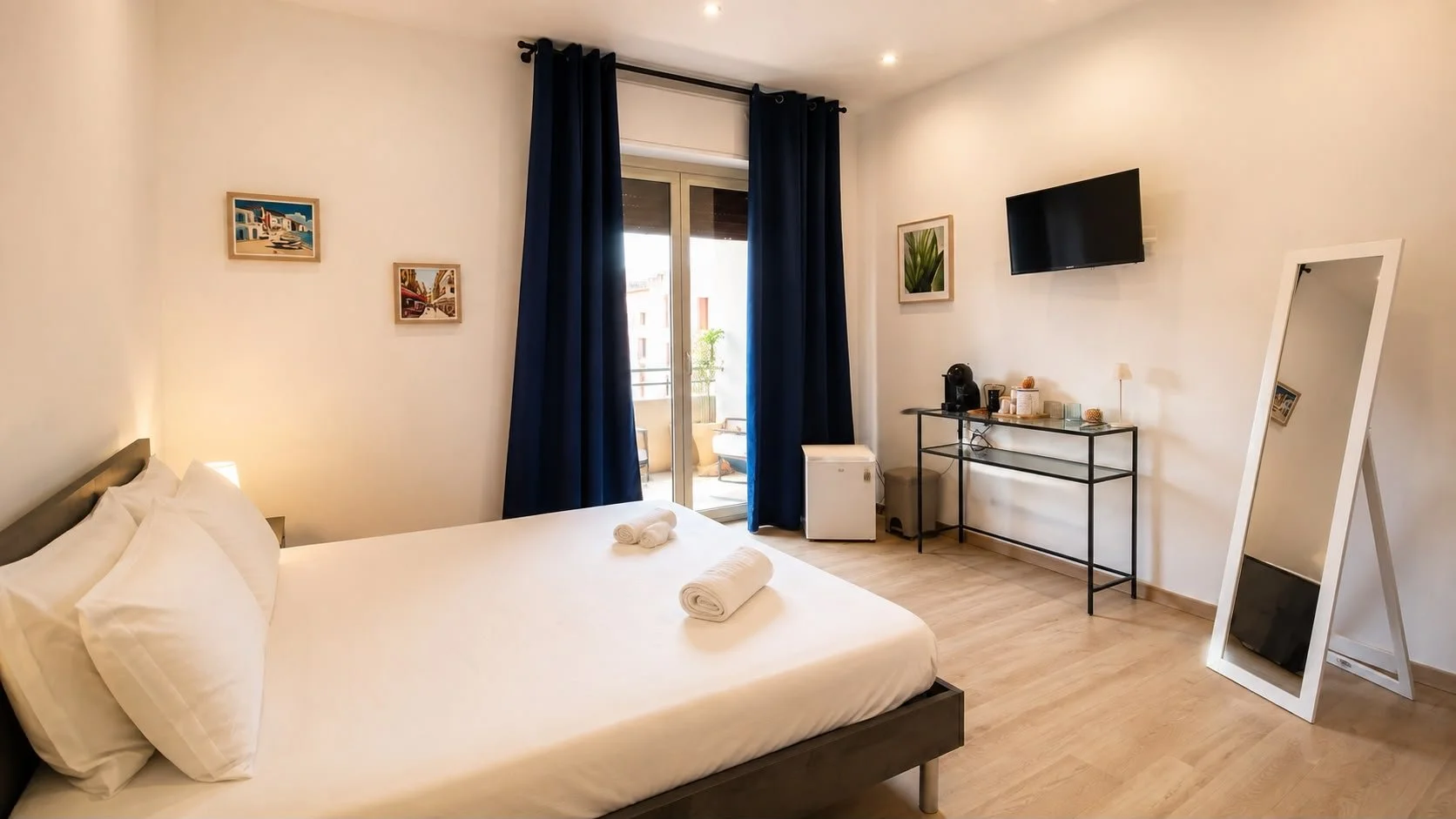 Camera con letto matrimoniale, specchio a figura intera, balcone con vista, pareti bianche decorate con quadri, lampada, televisore, aria condizionata, tavolo con caffettiera e snack.