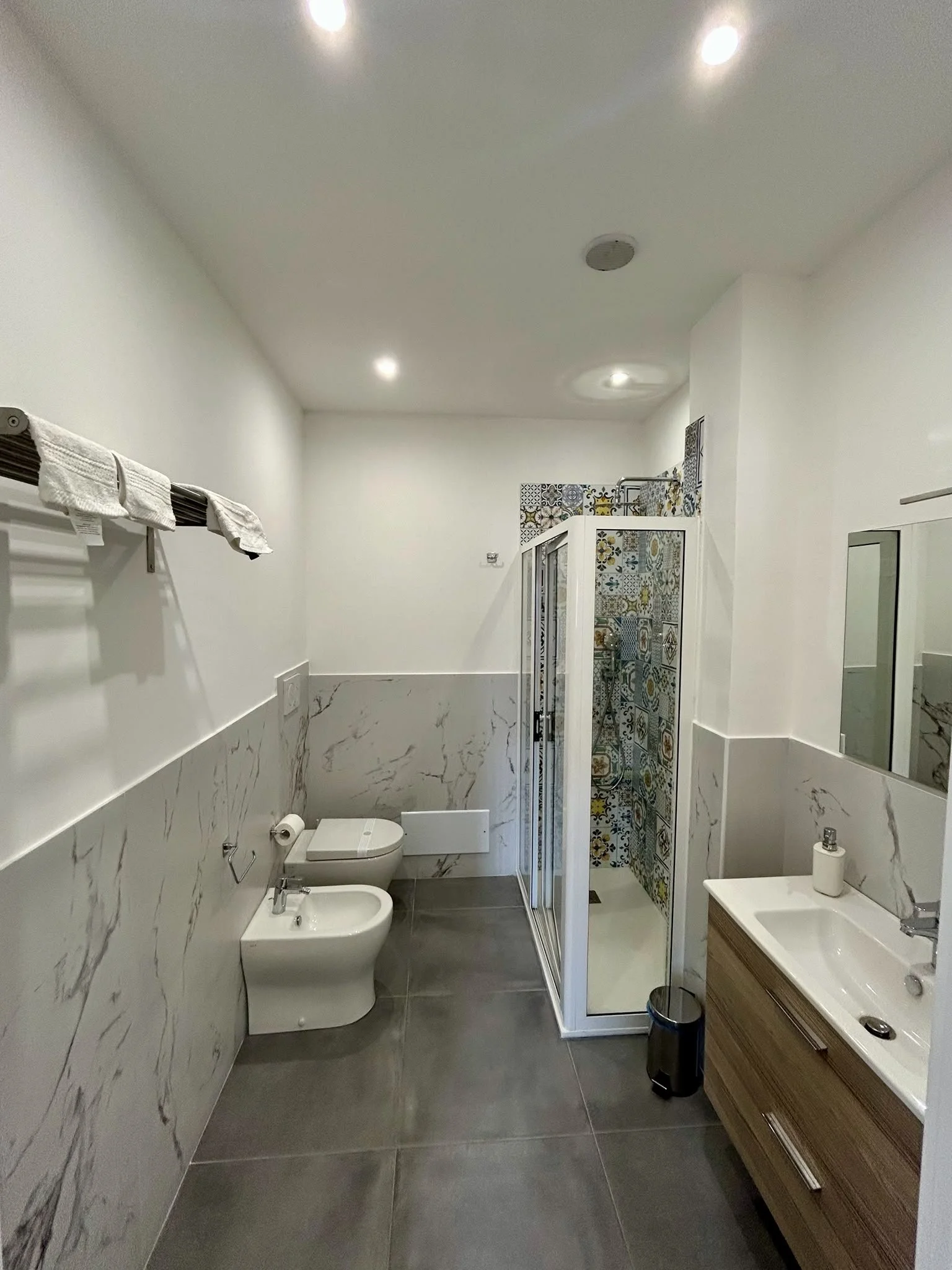 Bagno moderno con pareti in marmo e piastrelle decorative nella zona doccia, lavabo con mobile in legno e specchio, WC e bidet, asciugamani sulle mensole, finestra e luce a soffitto.
