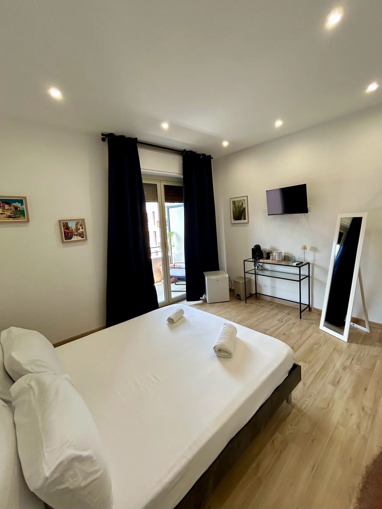 Una stanza con un letto con lenzuola bianche e due asciugamani arrotolati sopra. Sul muro ci sono quattro quadri e un televisore a schermo piatto. C'è una grande finestra con tende nere e un balcone.