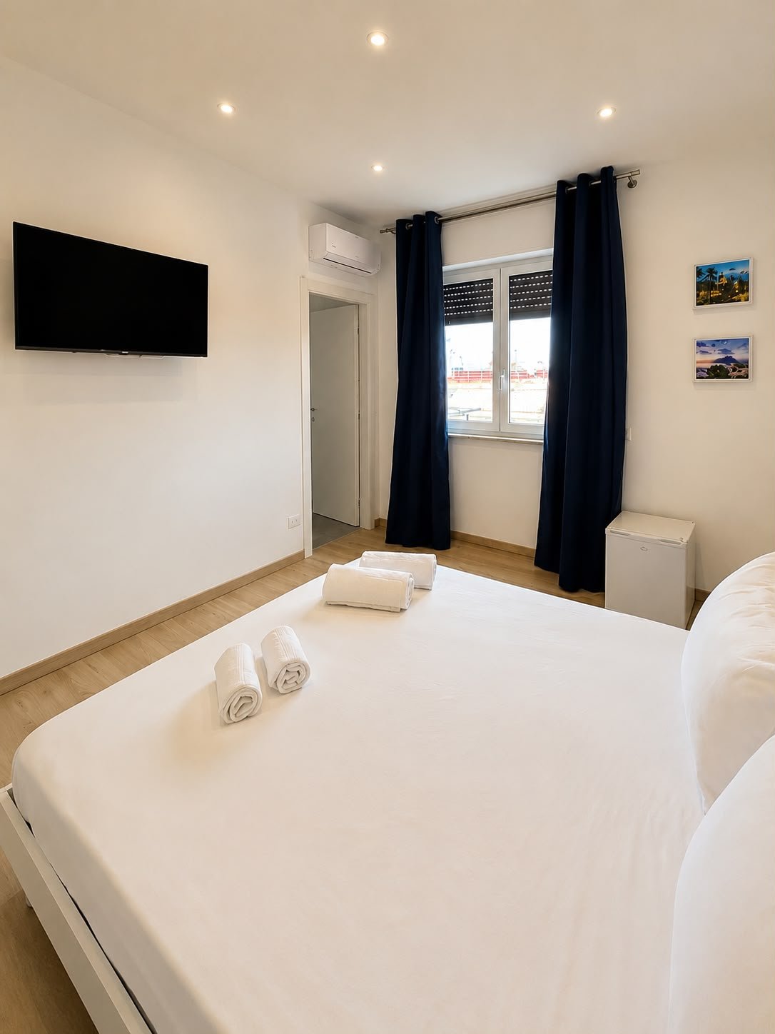 Camera da letto luminosa con letto bianco, tende blu, pareti bianche, televisore a schermo piatto, aria condizionata, fotografie alle pareti, mini frigorifero, e finestra che dà sulla città.