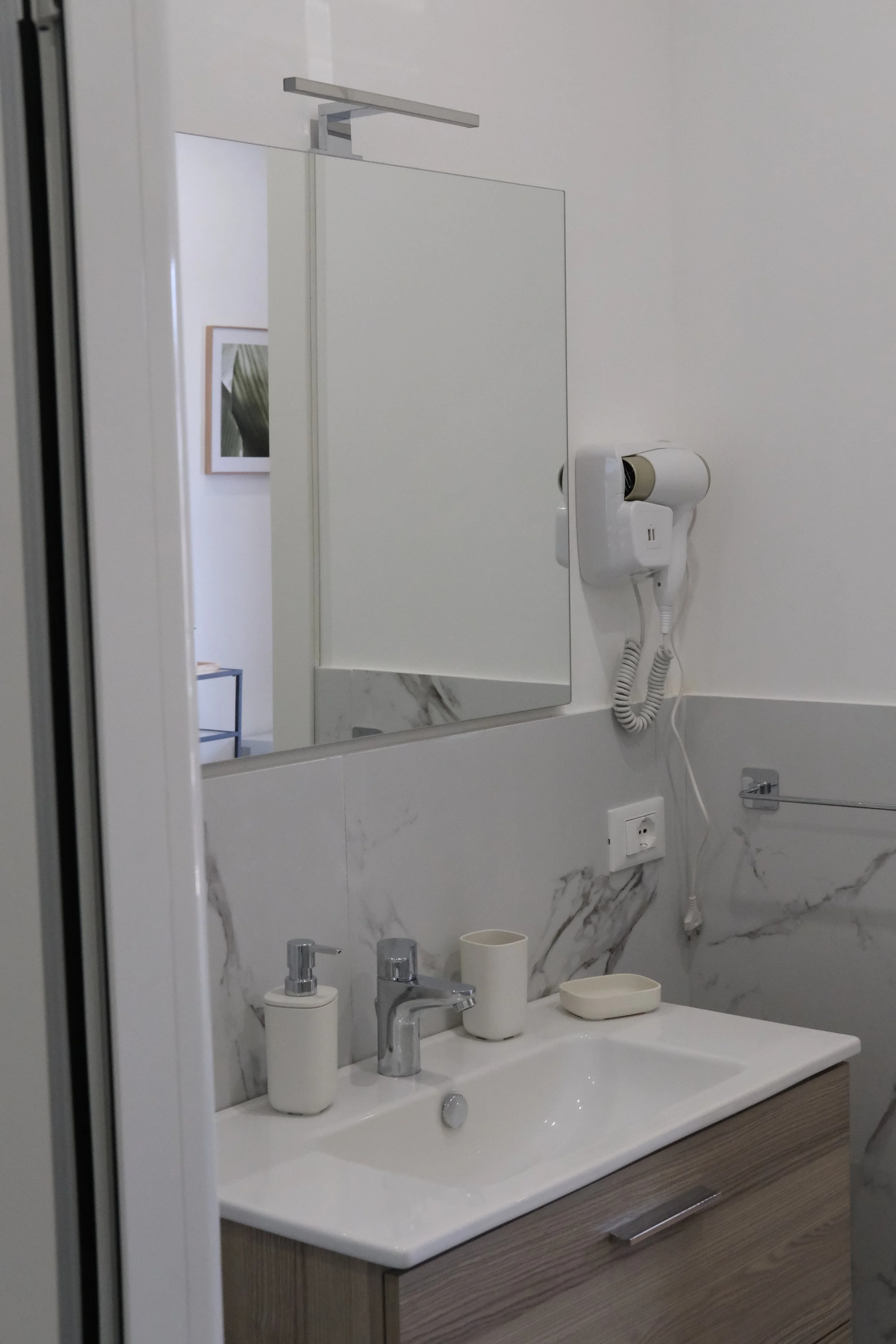 Specchio, lavabo con due dispense per sapone e una tazza, asciugamano di carta, specchio bar, presa di corrente, asciugacapelli attaccato al muro, pareti rivestite con lastre di marmo.