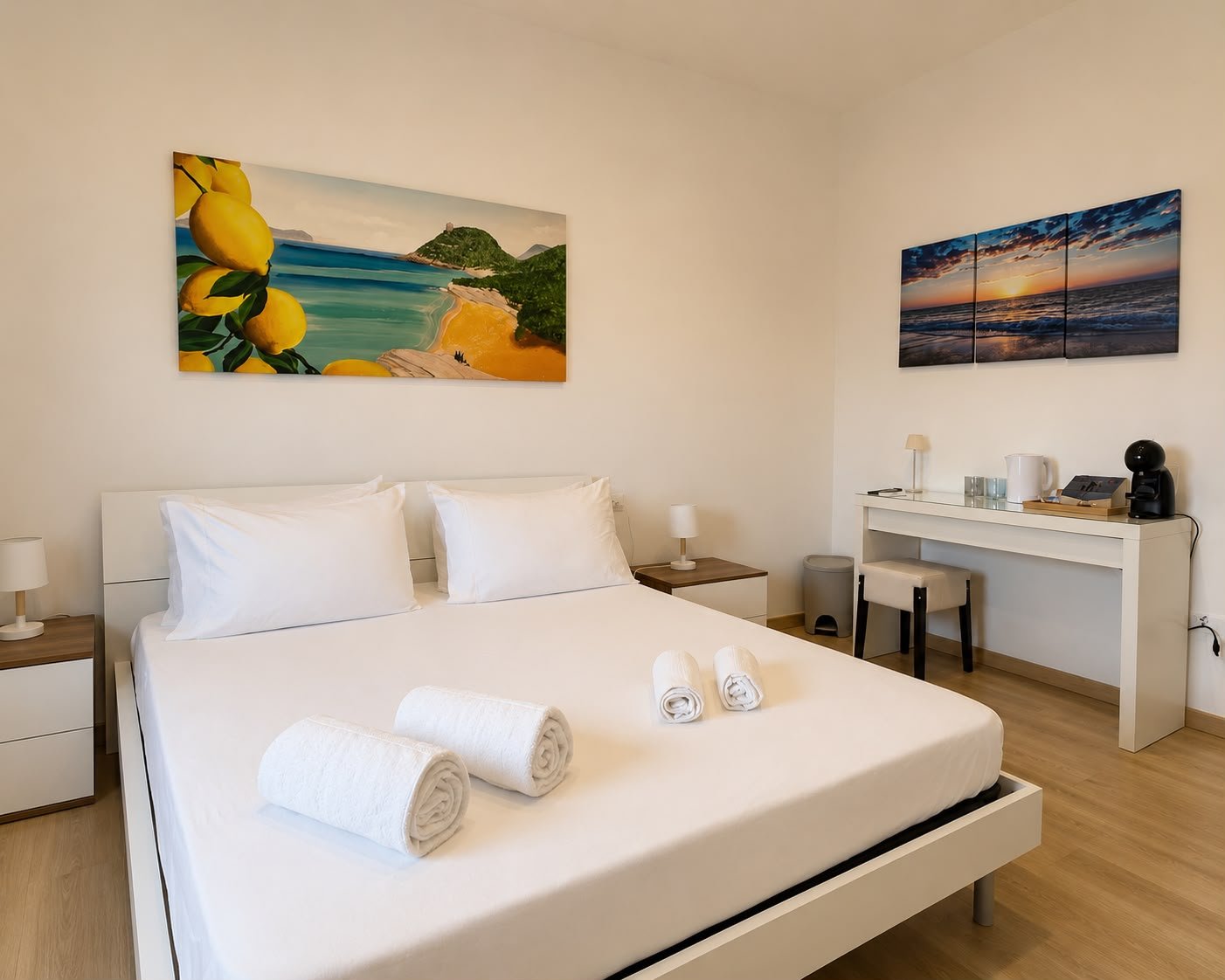 Camera da letto con letto matrimoniale bianco, due cassettiere con lampade ai lati, quadri con paesaggi marini e costieri, asciugamani arrotolati sul letto, scrivania con tazza, macchina da caffè e altri oggetti, sedia e lampada sullo sfondo.
