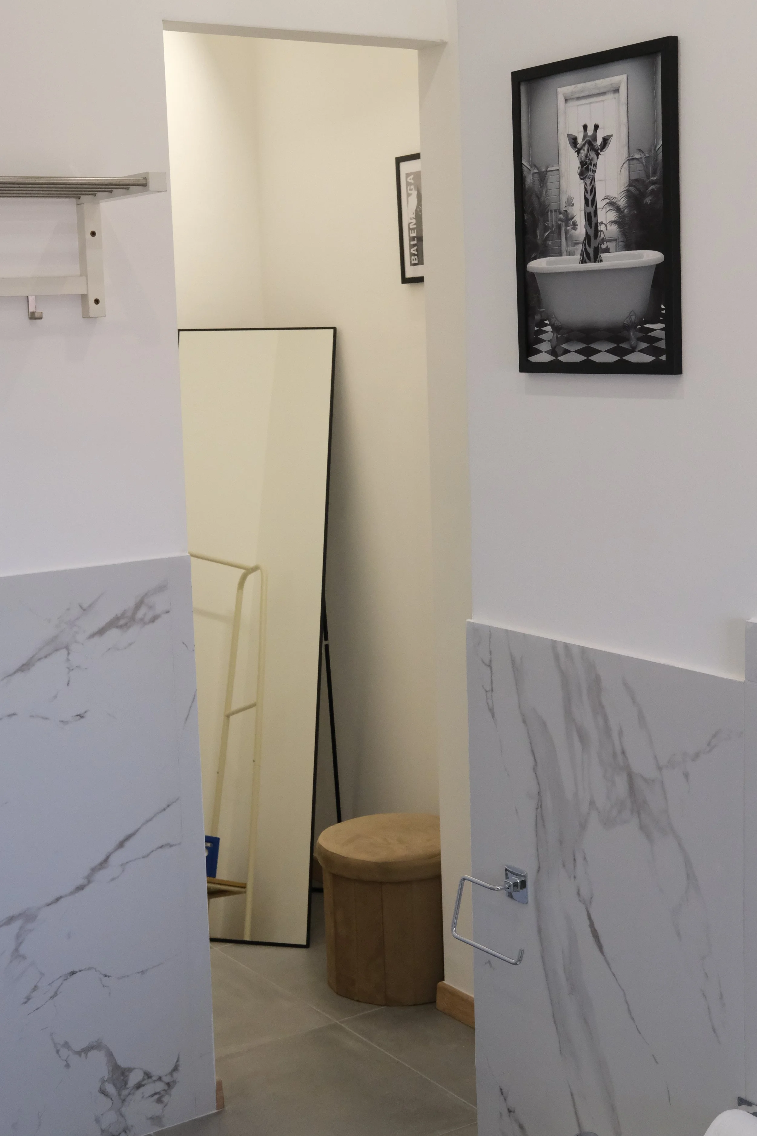 Specchio a terra riflette un ambiente con cornice nera, un quadro con giraffa in vasca da bagno e un tavolo di legno, pareti bianche con decorazioni in marmo.