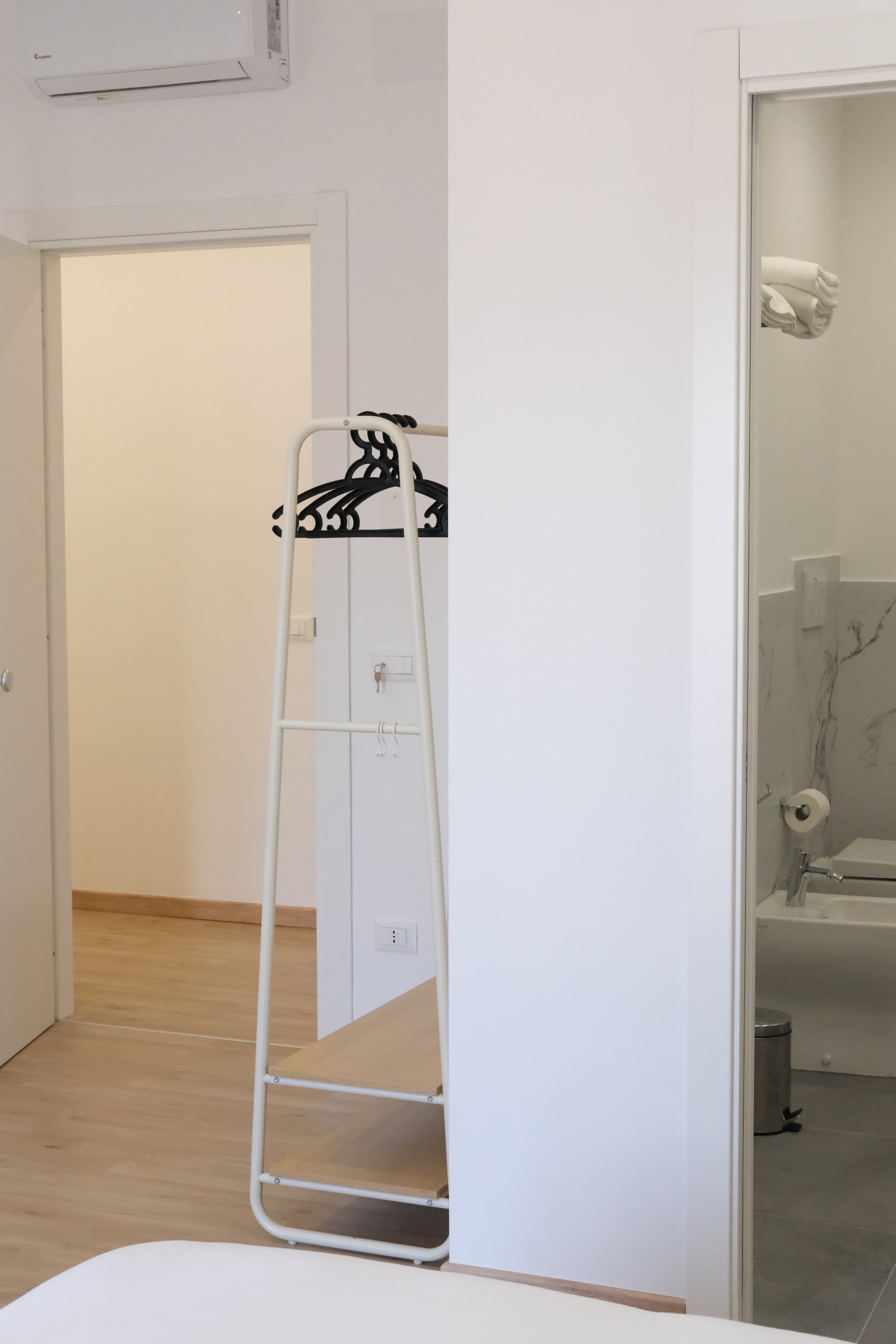 Una stanza con pavimento in legno chiaro, pareti bianche e una porta che dà su un bagno con lavabo e WC. Sulla sinistra si vede un dispositivo di climatizzazione a parete e un'appendiabiti nero con grucce.