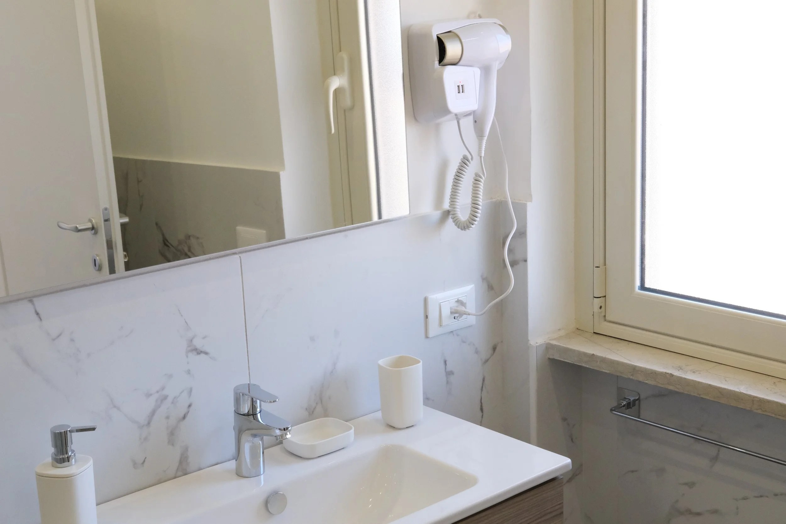 Specchio sopra lavabo con dispenser di sapone, tazza e porta spazzolini, finestra con cornice, asciugamani di carta e presa elettrica.