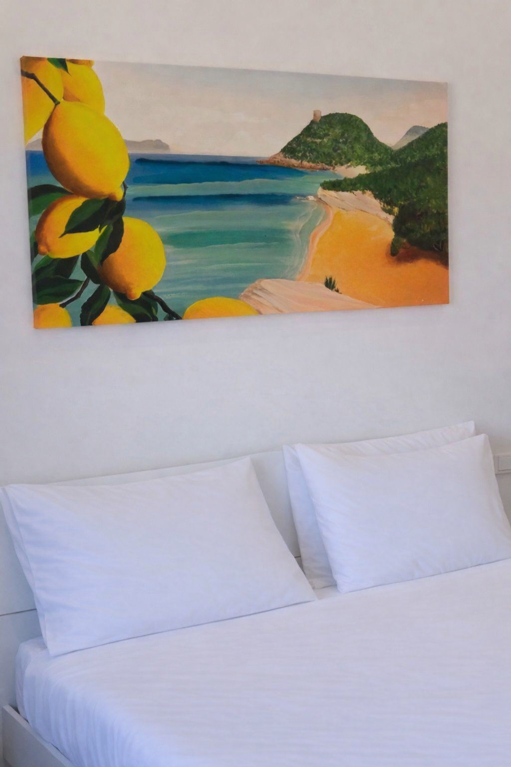 Un dipinto di una spiaggia con mare, colline verdi e limoni in primo piano appeso sopra un letto con cuscini bianchi.
