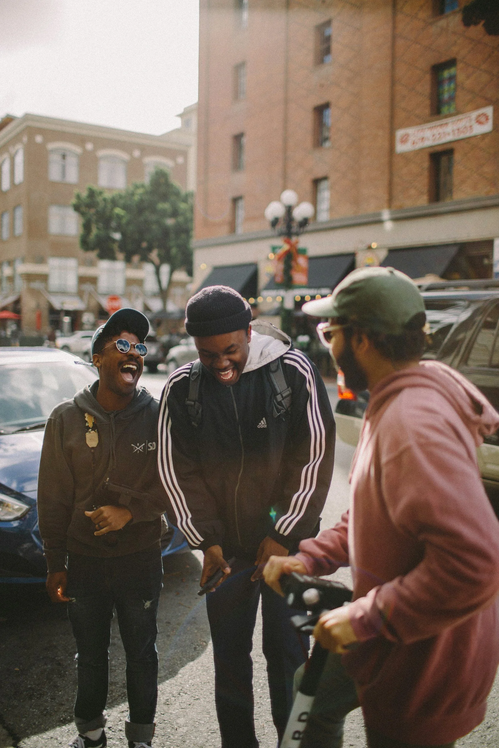 Three young men laughing and talking on a city street during daytime. They are dressed casually with backpacks and hats, standing in front of parked cars, and buildings with storefronts and a streetlamp are visible in the background.