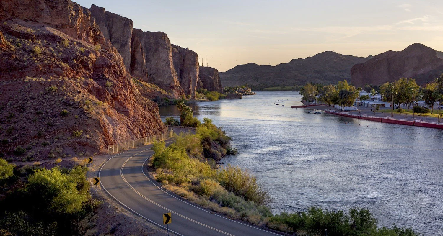 A winding river with a road running alongside it, surrounded by hillside on the left with large rocks and sparse vegetation, and a small town with trees on the right Bank, mountains in the background, and the sky at sunset or sunrise.