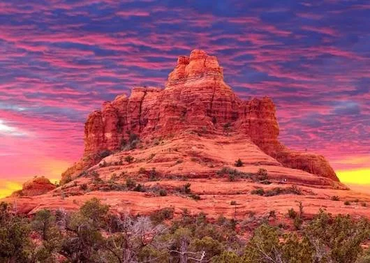 A large reddish-brown rock formation resembling a fortress or castle under a colorful sunset sky with pink, purple, and yellow hues, and sparse desert vegetation at the base.