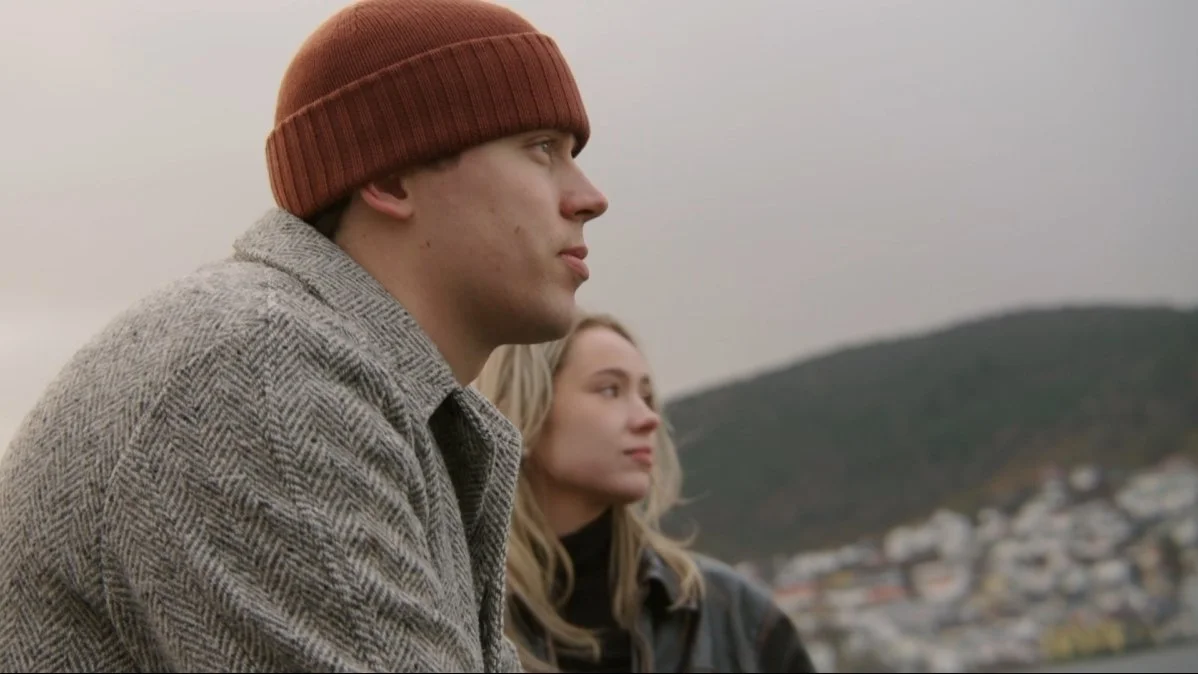 En mann og en kvinne med havet og fjell i bakgrunnen, ser ut til å tenke eller vente.