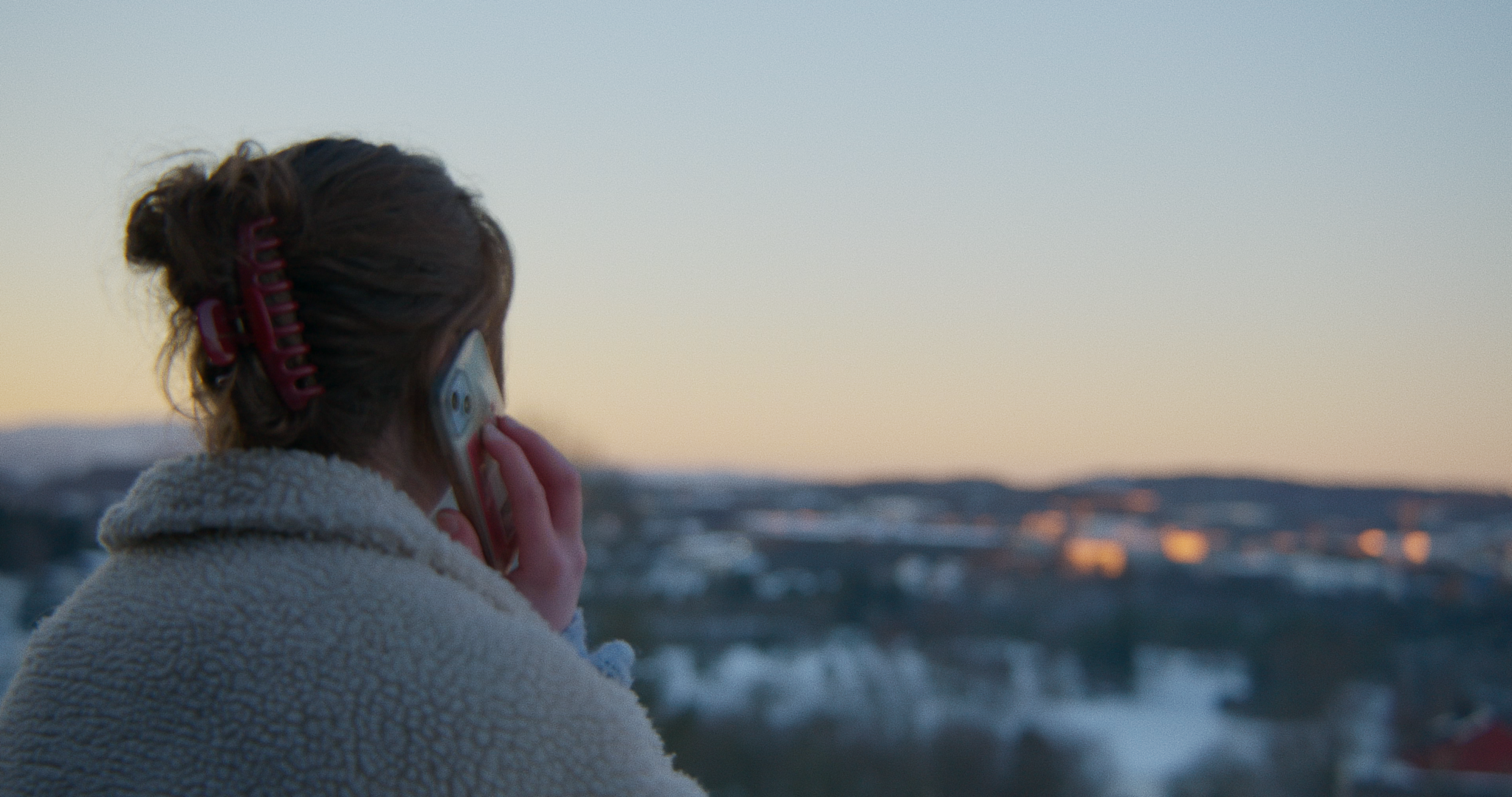 Person med briller som snakker i telefon, med hårbøyle, ute i landskapet under solnedgang.