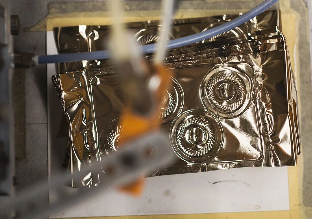 Top-down view of a reflective metal mold or baking pan with three circular cavities, placed on a work surface with wires and tools nearby.