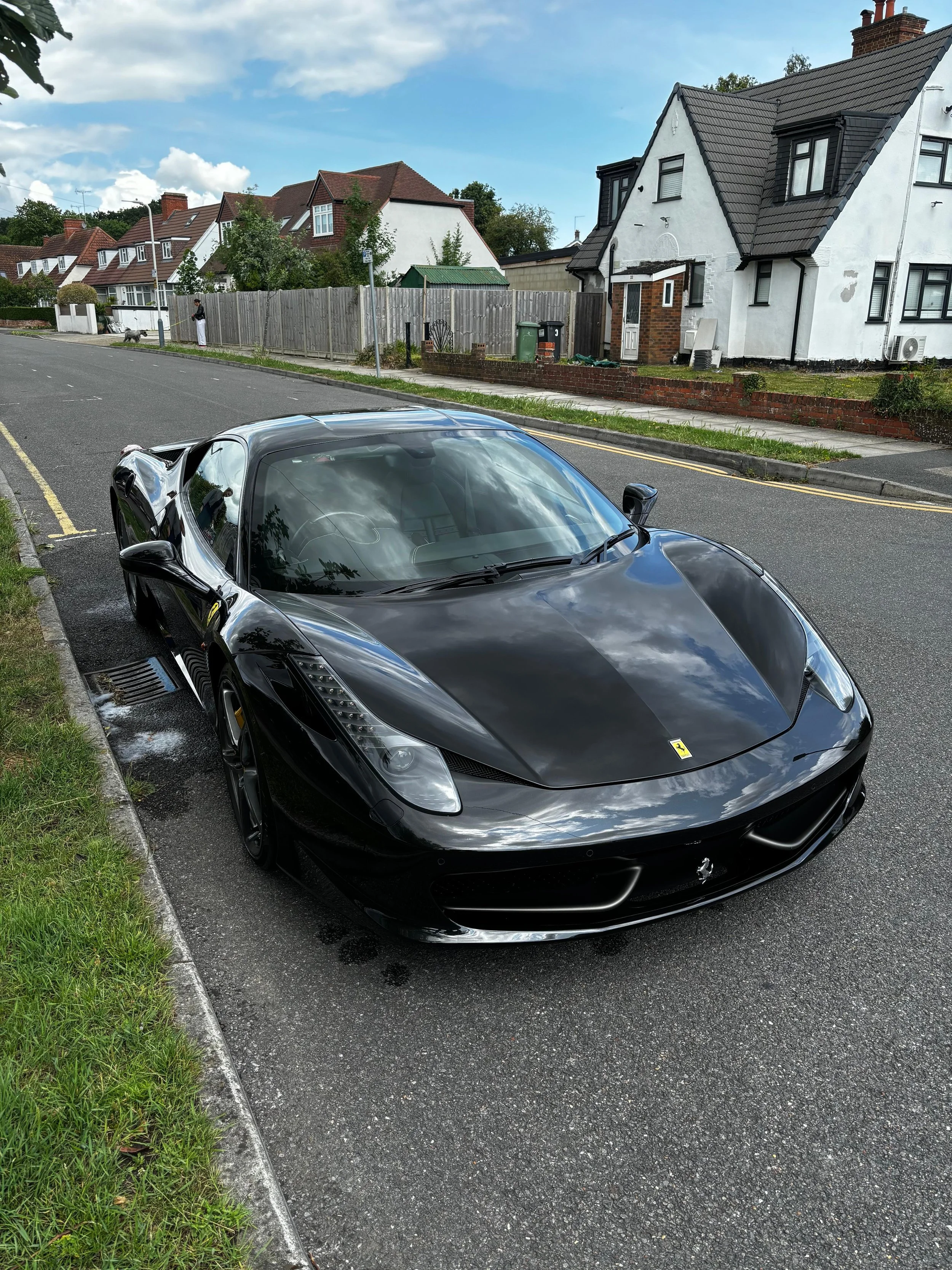 A shiny black Ferrari sports car parked on the street with houses and a sidewalk in the background.