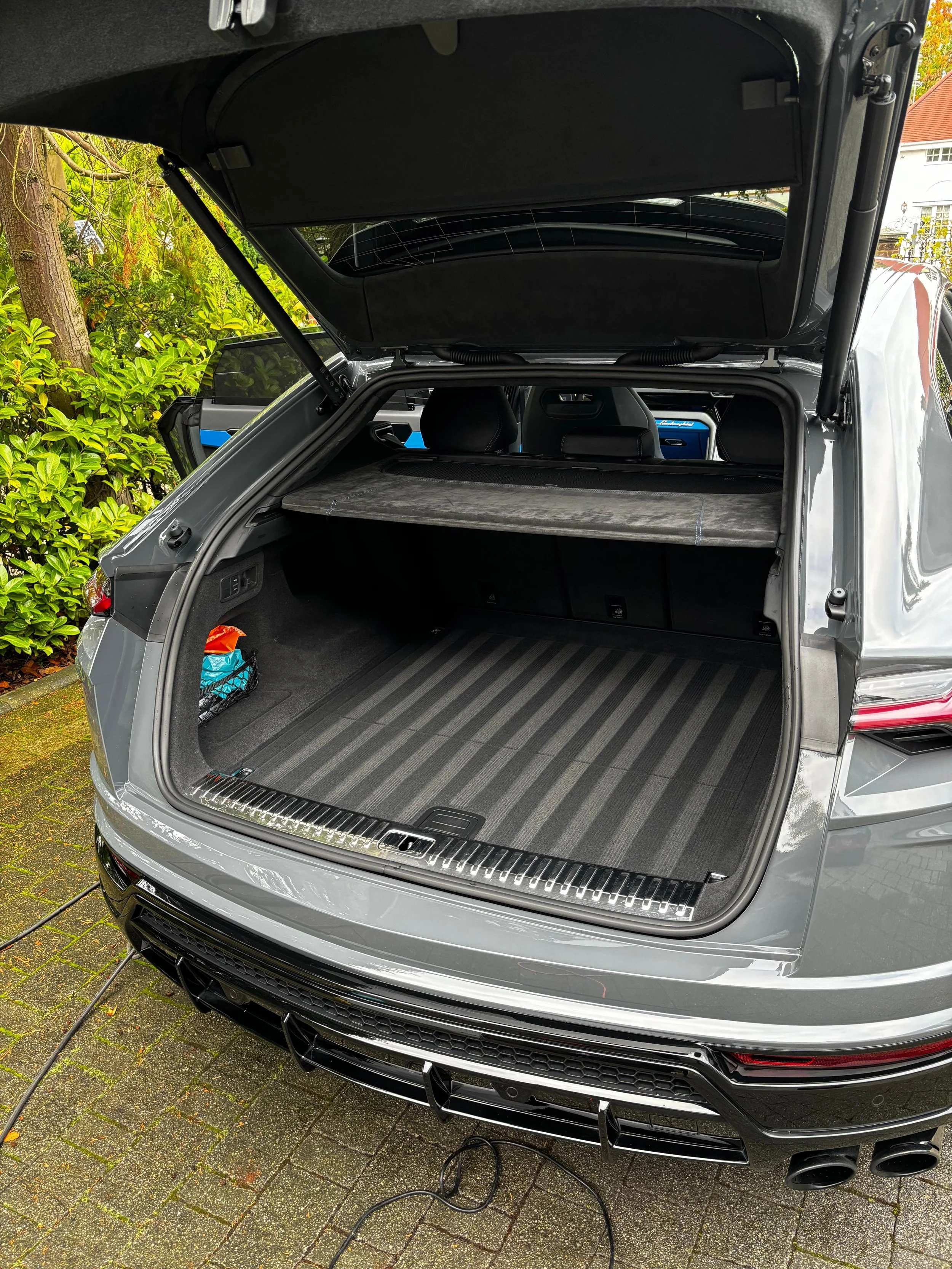 The back of a silver luxury SUV with its trunk open, showing an interior cargo space with a dashboard cover, on a brick driveway near green bushes and a house.