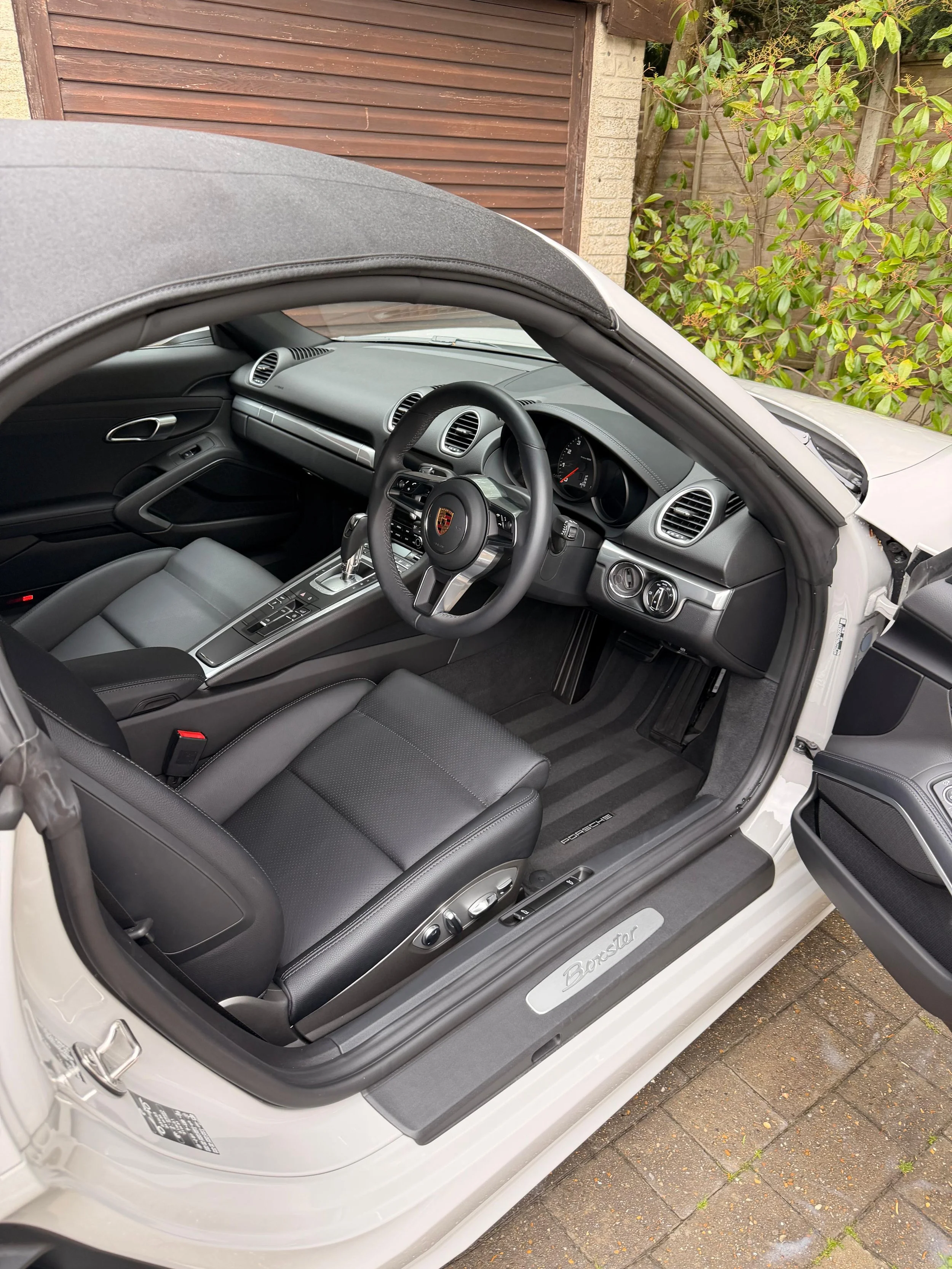 Interior of a Porsche Boxster sports car with black leather seats and modern dashboard, viewed from the driver's side with the door open, parked on a brick surface with greenery and a wooden fence in the background.