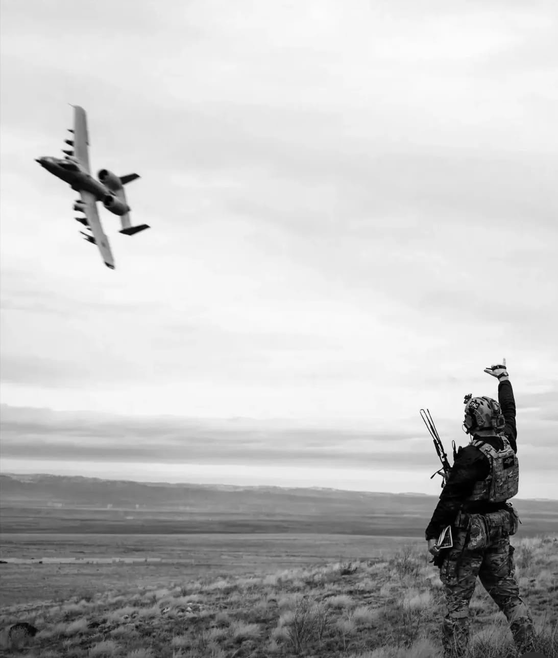 A soldier in military gear standing on a grassy plain, signaling with his right arm while watching a fighter jet flying overhead in a cloudy sky.