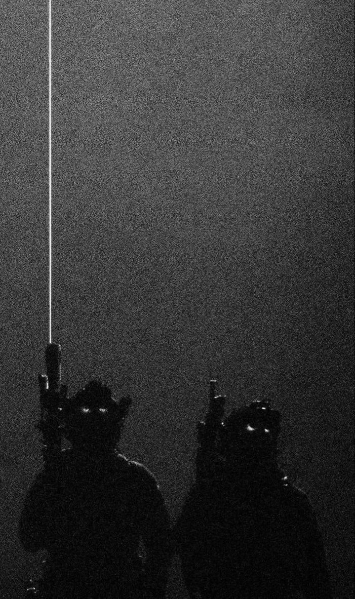 Two soldiers in tactical gear with night vision goggles, standing in darkness.