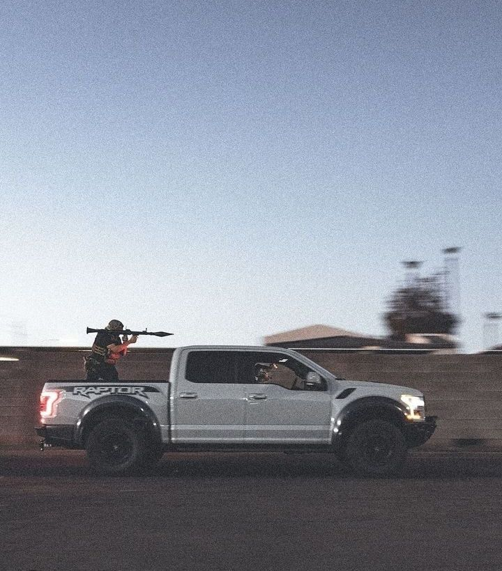 A silver pickup truck with black wheels and the word 'Raptor' on the side driving on a road at dusk. A person in the truck's bed is holding a long rifle, aiming it into the distance.