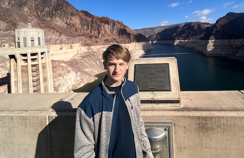 Logan Galdabini standing on a dam overlooking a large reservoir with rocky canyon walls in the background. The boy is wearing a blue and gray jacket and casting a shadow on the concrete surface.