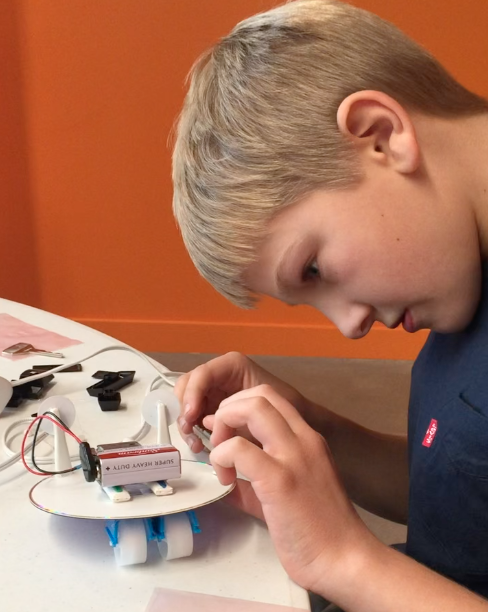 Young boy working on a small robotics project with electronic components and wires on a table.