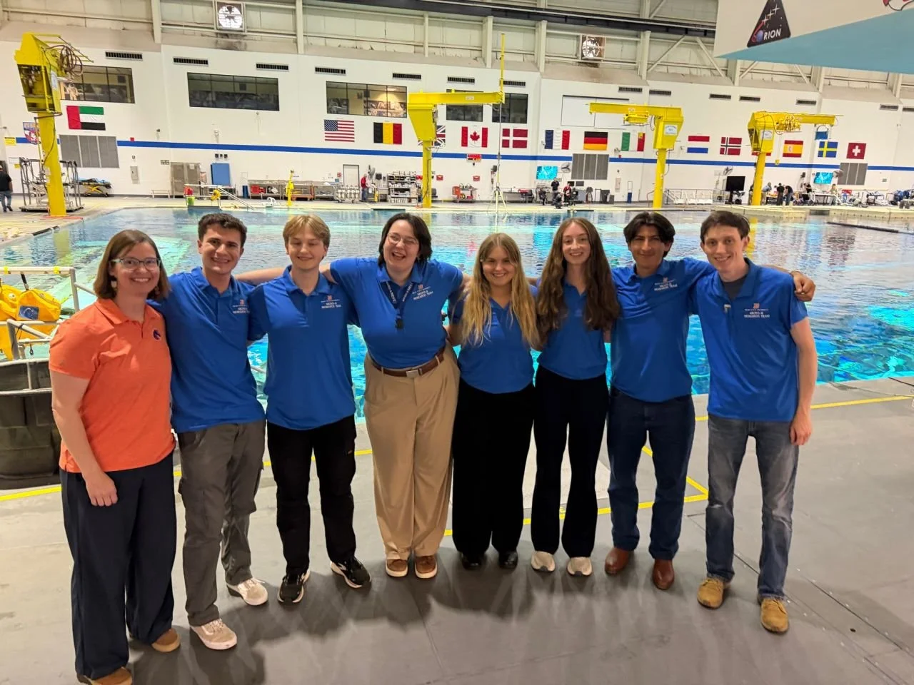 Group of seven people standing side by side in front of an indoor swimming pool, smiling at the camera. The pool has diving boards and flags from different countries hanging above.