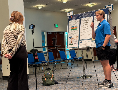 Logan Galdabini at the Conference on Undergraduate Research. Young woman filming a presentation on a poster board in a conference room with empty blue chairs.